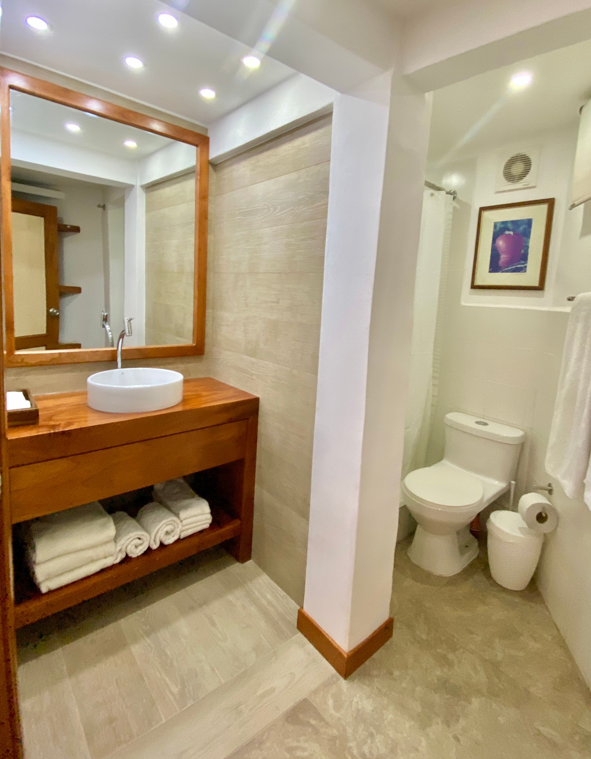 Bathroom with a wooden vanity, white sink, and toilet. Beige walls, white towels, and a framed picture add color.