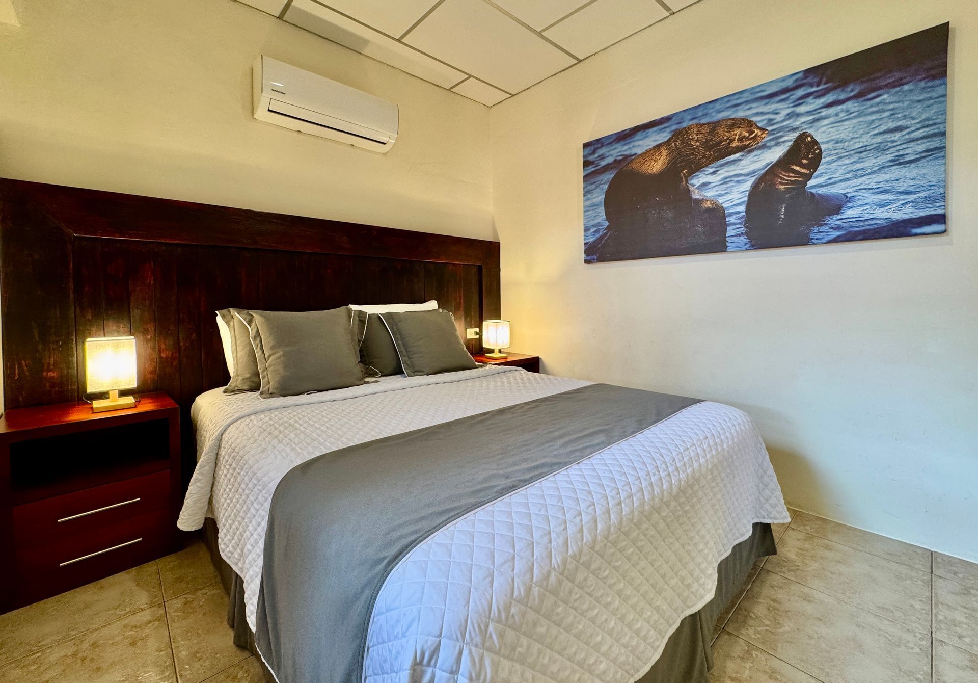 Bedroom with a dark wood headboard, a white and gray bedspread, bedside tables with lamps, and a sea lion artwork.