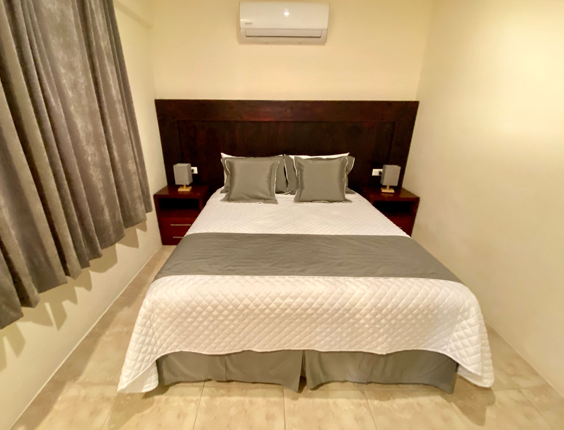 A hotel bedroom with a bed, two nightstands, and an air conditioning unit. The bed has gray and white bedding.