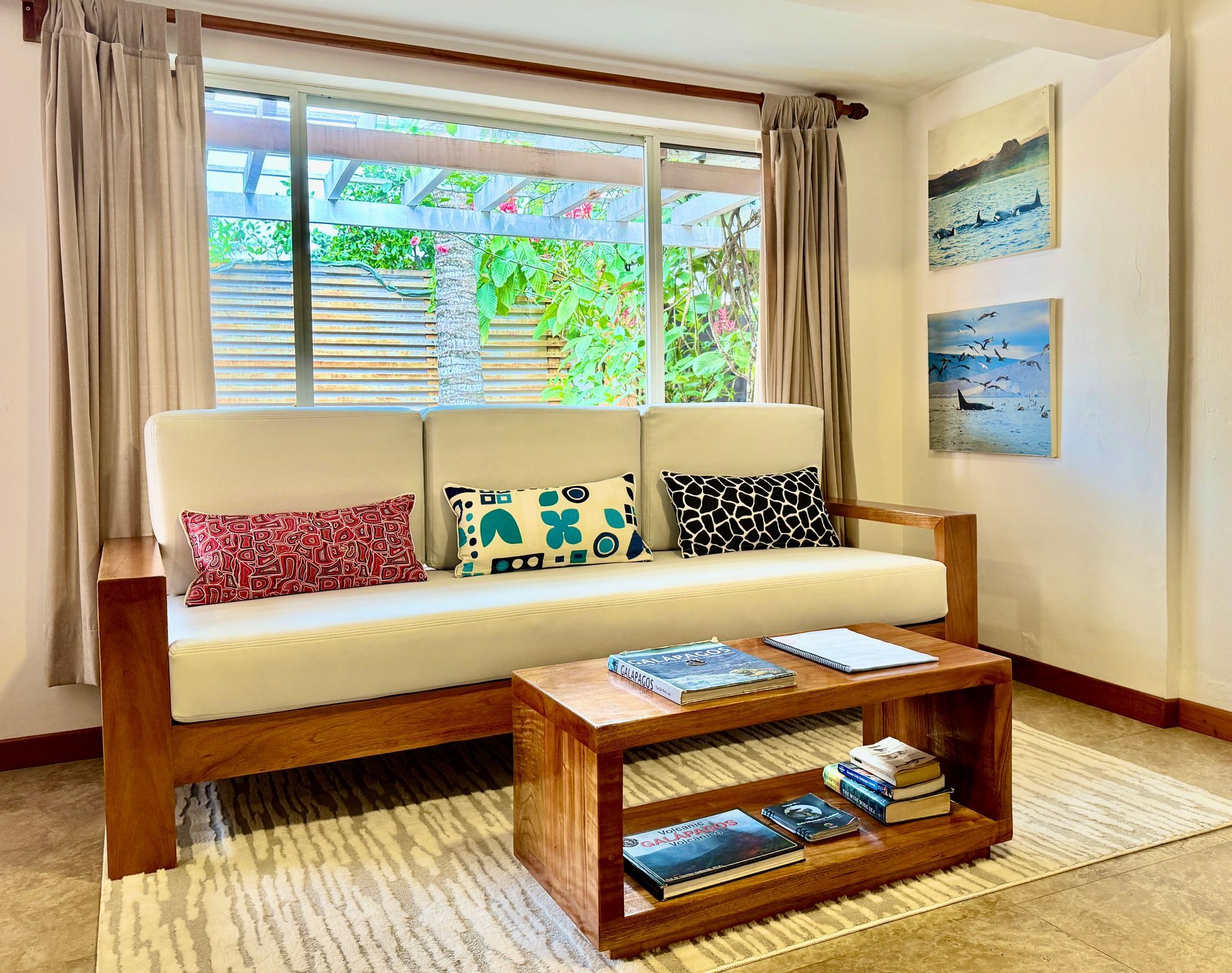A living room with a wooden couch, coffee table, and window overlooking greenery. Pillows and art decorate the space.