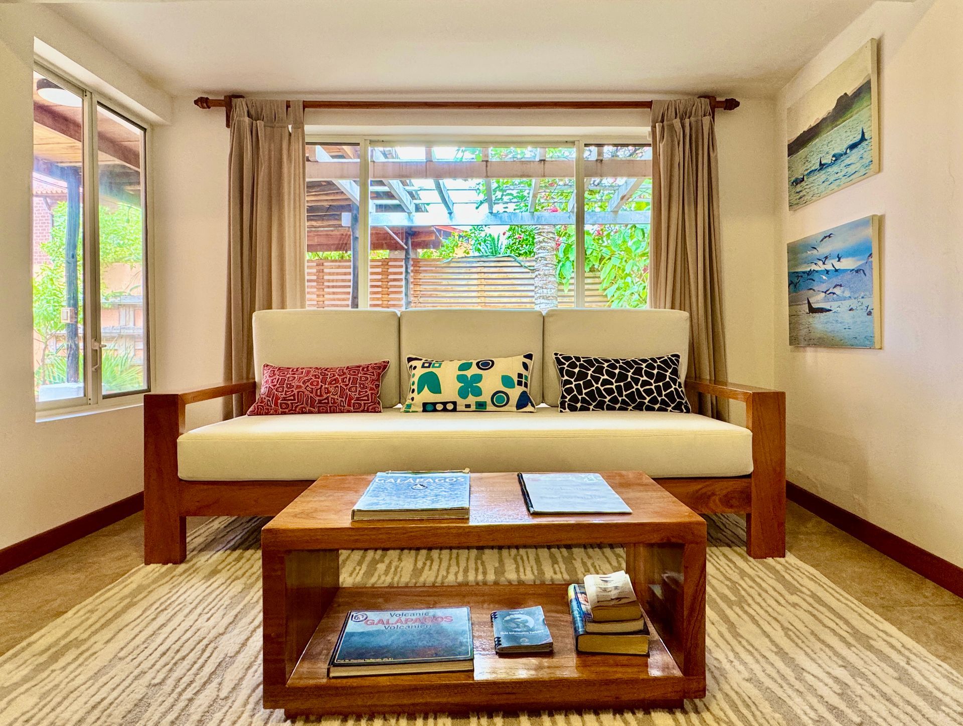 Living room with cream-colored sofa, wooden coffee table, and artwork on the wall. A large window looks out onto greenery.