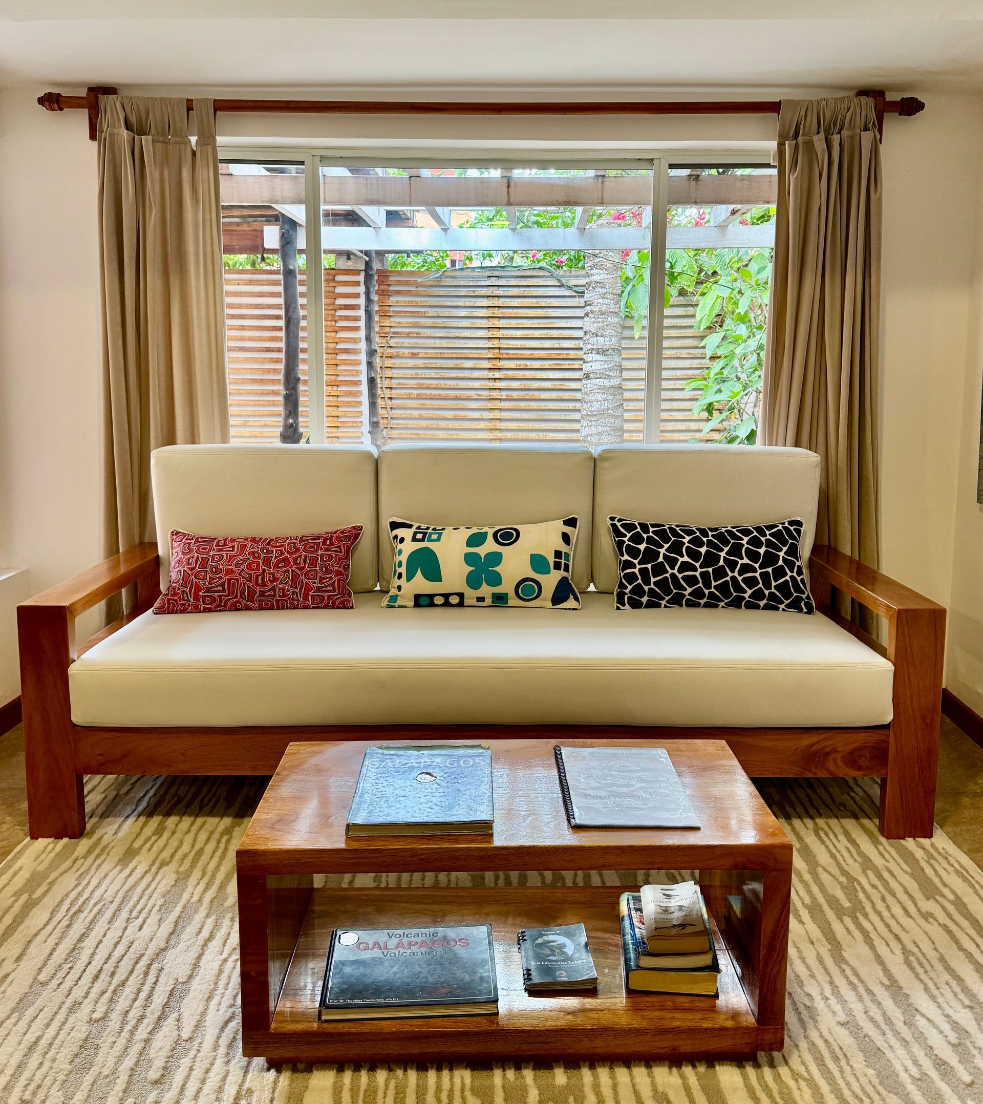 Living room with a wooden-framed sofa, coffee table, and window overlooking a garden. Cushions with patterned designs.