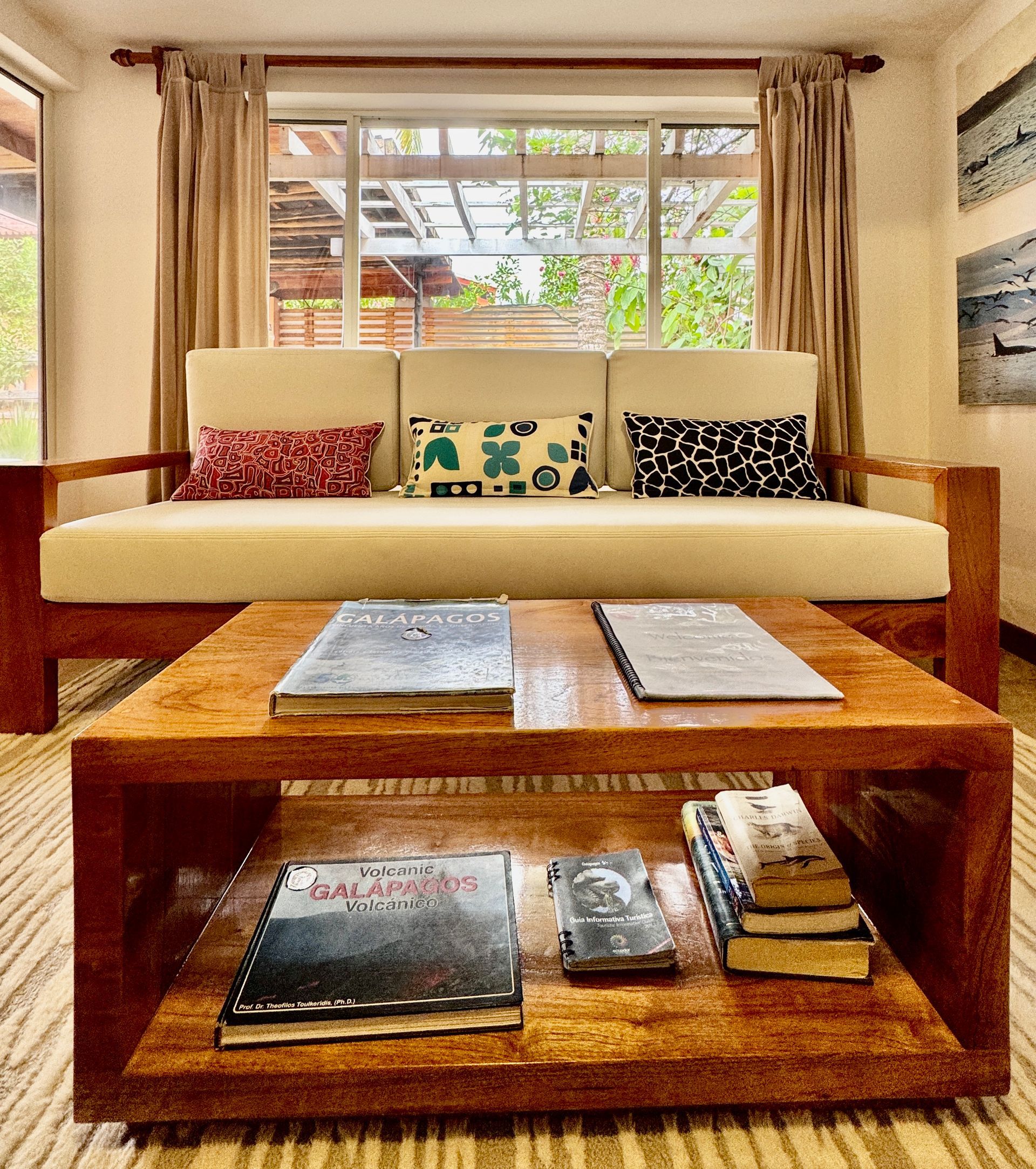 Living room with a wooden sofa and coffee table holding books, facing a window. Beige and brown tones dominate the scene.