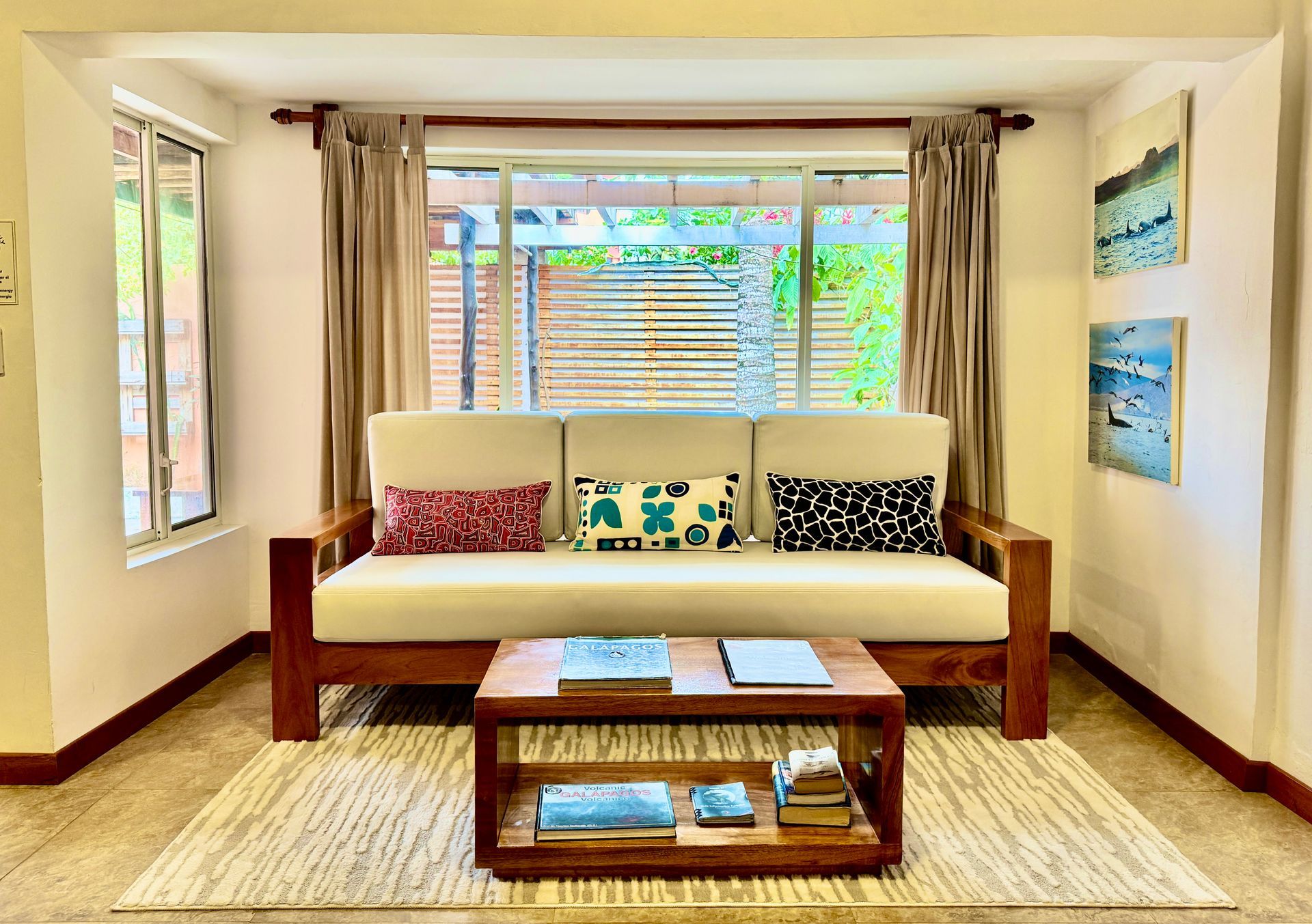 Living room with wooden sofa, coffee table, and patterned rug. Windows with beige curtains and artwork on the walls.