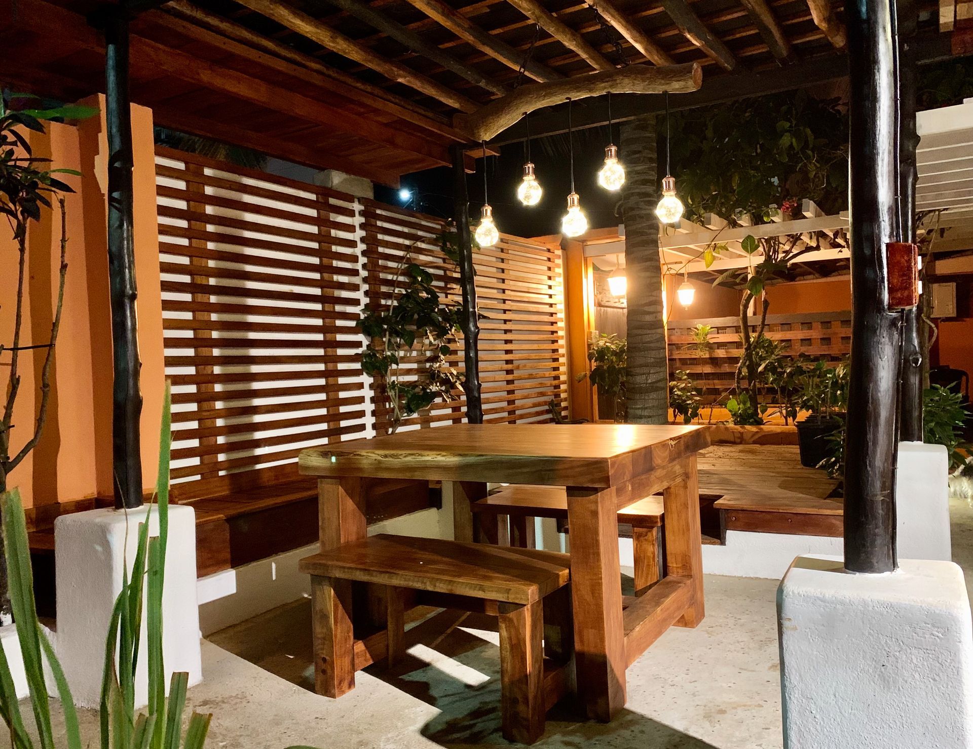 Outdoor dining area with wooden table and benches, overhead lighting, and slatted wood wall.