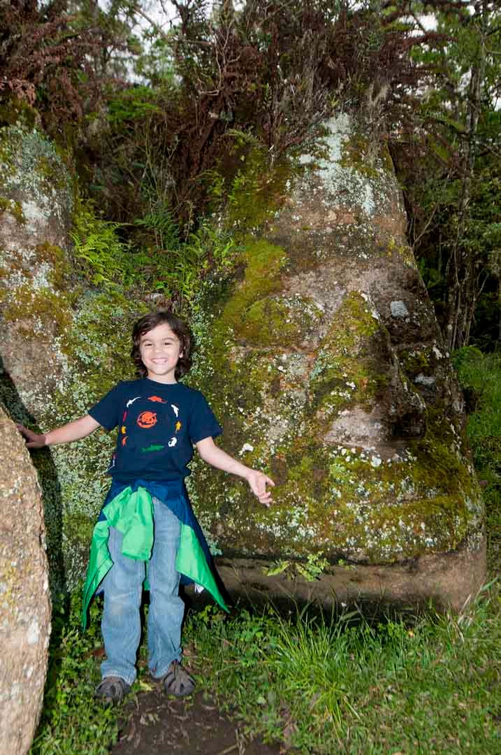 A young boy is standing next to a large rock in the woods.