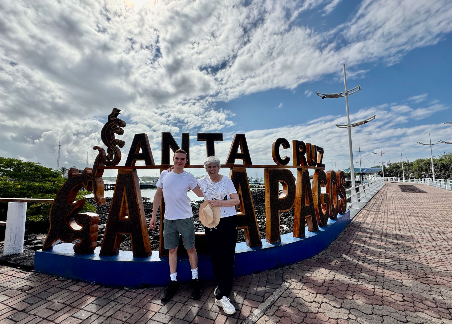Two people posing by the Santa Cruz Galapagos sign under a bright cloudy sky.