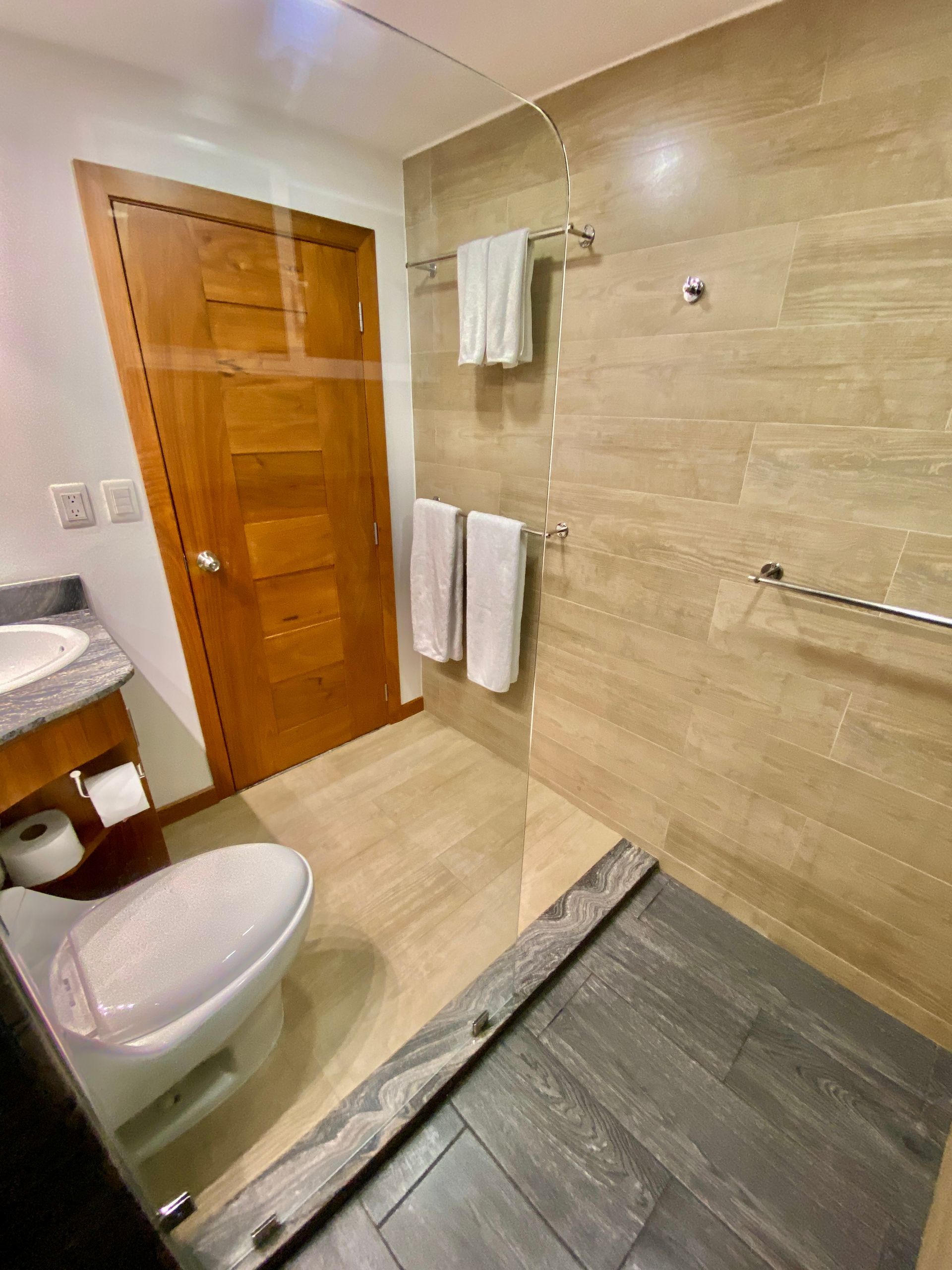 Bathroom with beige tiled walls and floor, wooden door, and glass shower enclosure. Two white towels hang on the wall.