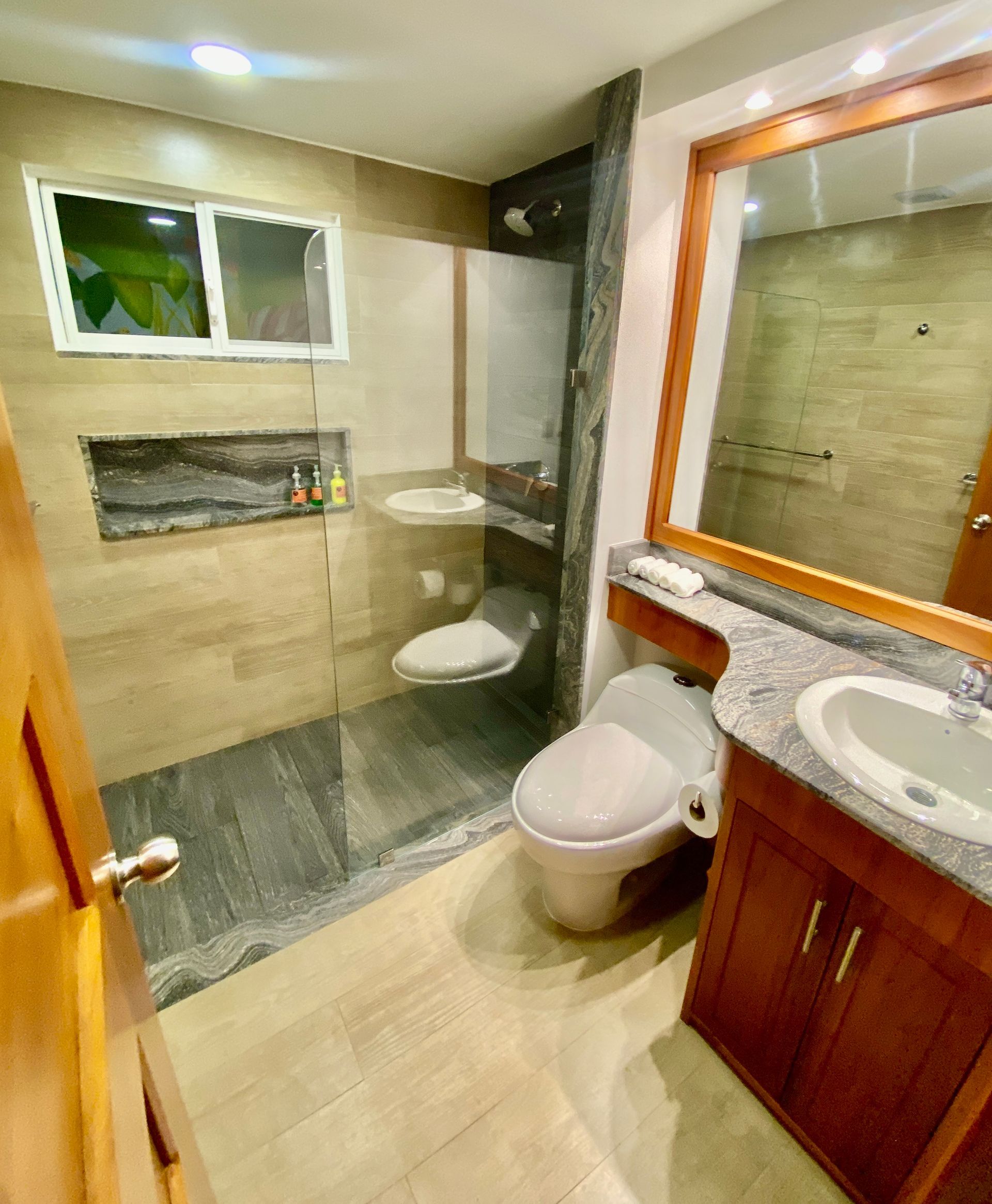 Bathroom with a walk-in shower, toilet, vanity, and a window. The walls are beige and gray tile, with wooden cabinetry.