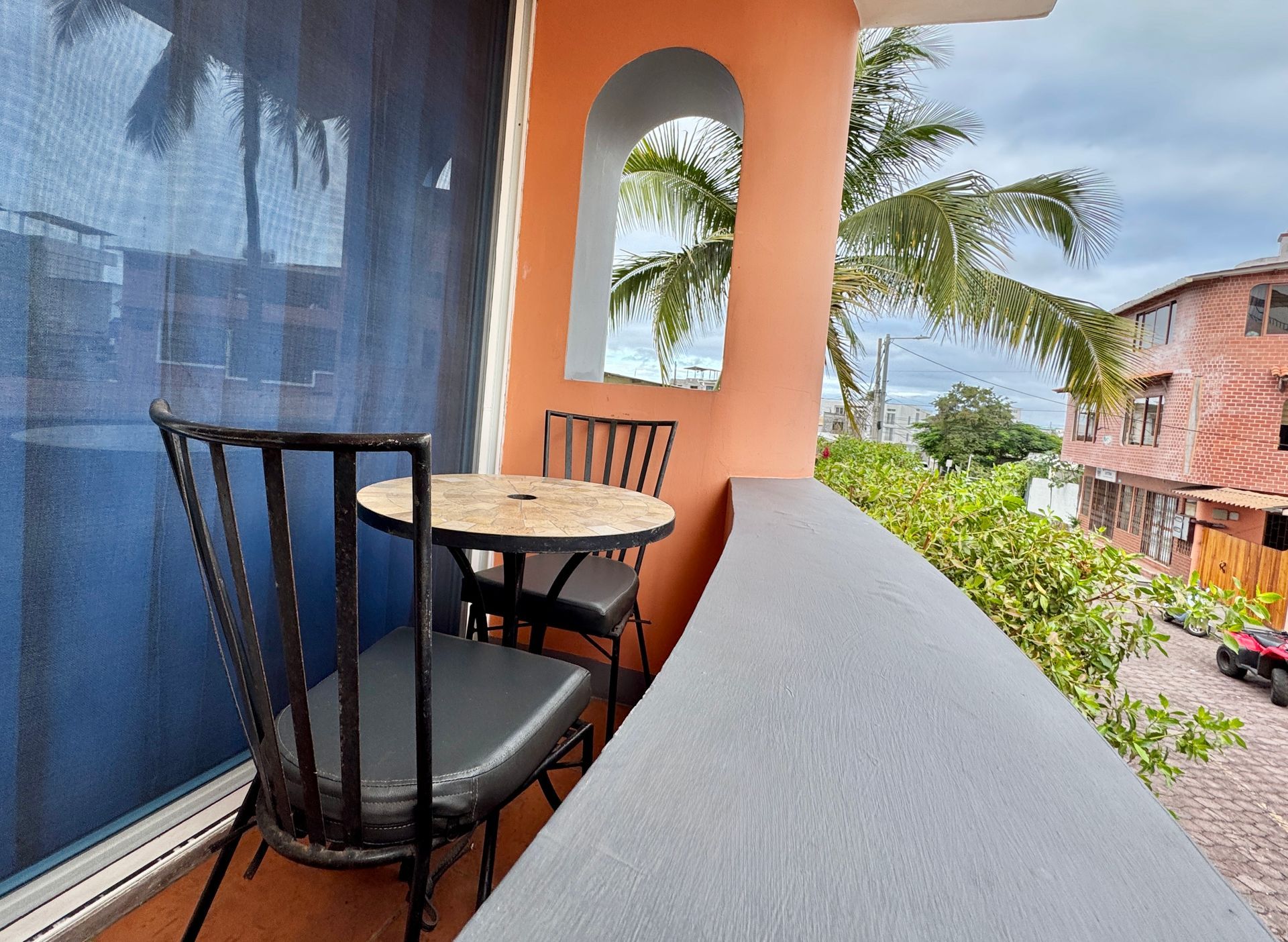 Balcony with a small table and two chairs overlooking palm trees and the ocean, with blue shades on the left.