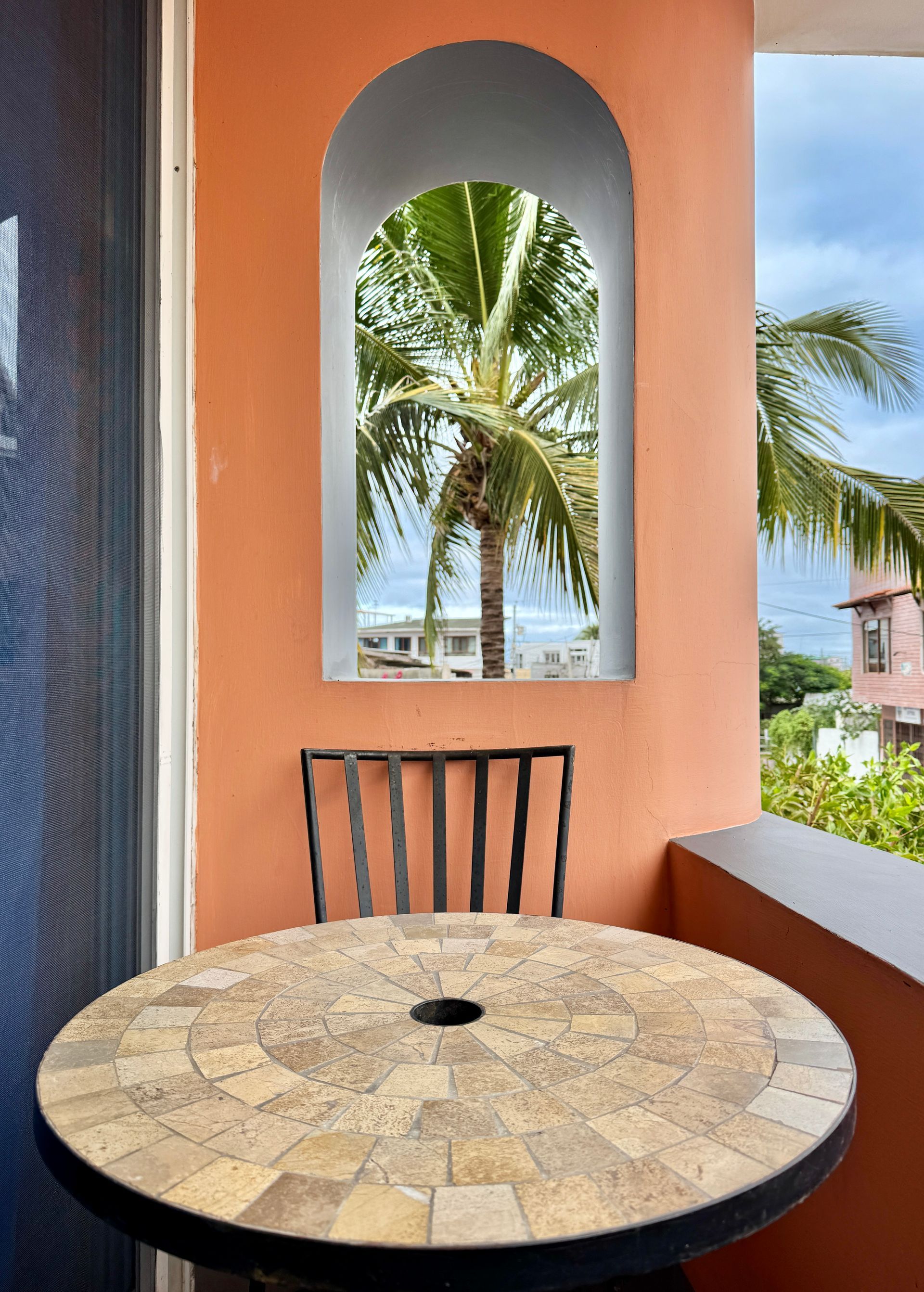 A small balcony with a mosaic-tiled table and a black metal chair. An arched window frames a palm tree and ocean view.