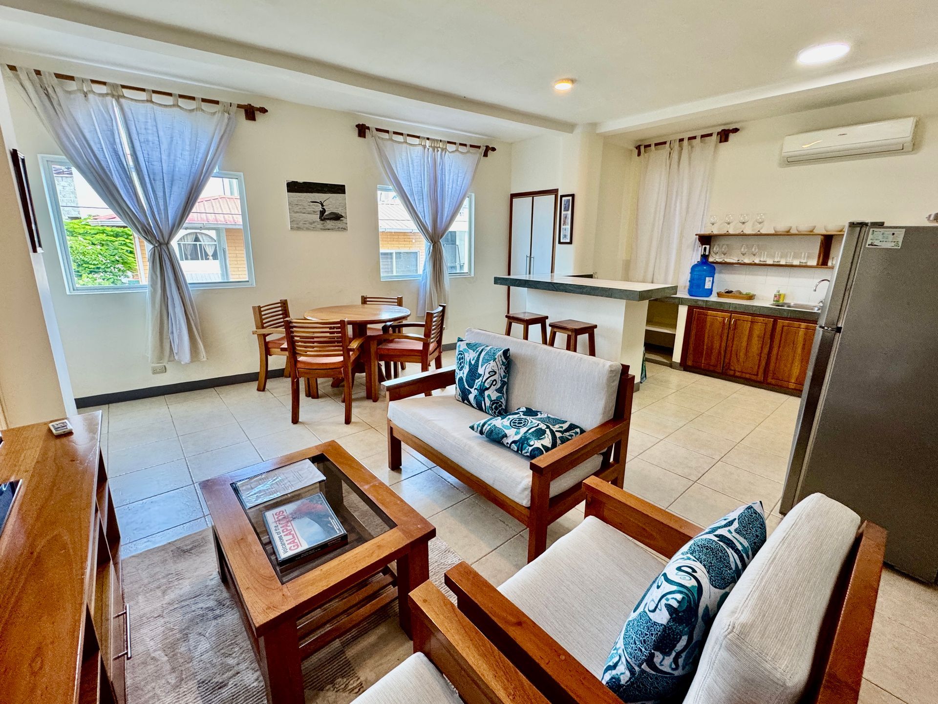 Living room with wooden furniture, dining table, and open kitchen area. Light-colored walls and tile flooring.