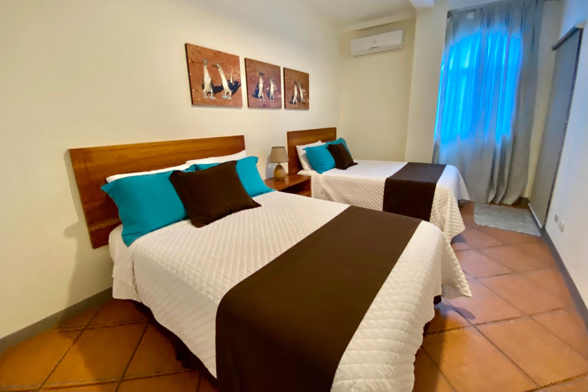 Two twin beds in a room with turquoise and brown accents, artwork, and a window with blue curtains.