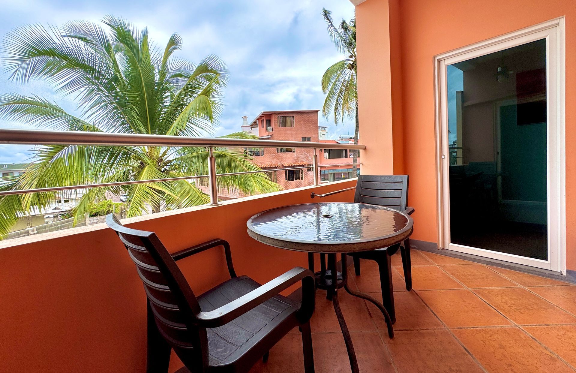 Balcony with table and chairs, overlooking palm trees and buildings. Bright orange walls, glass door.