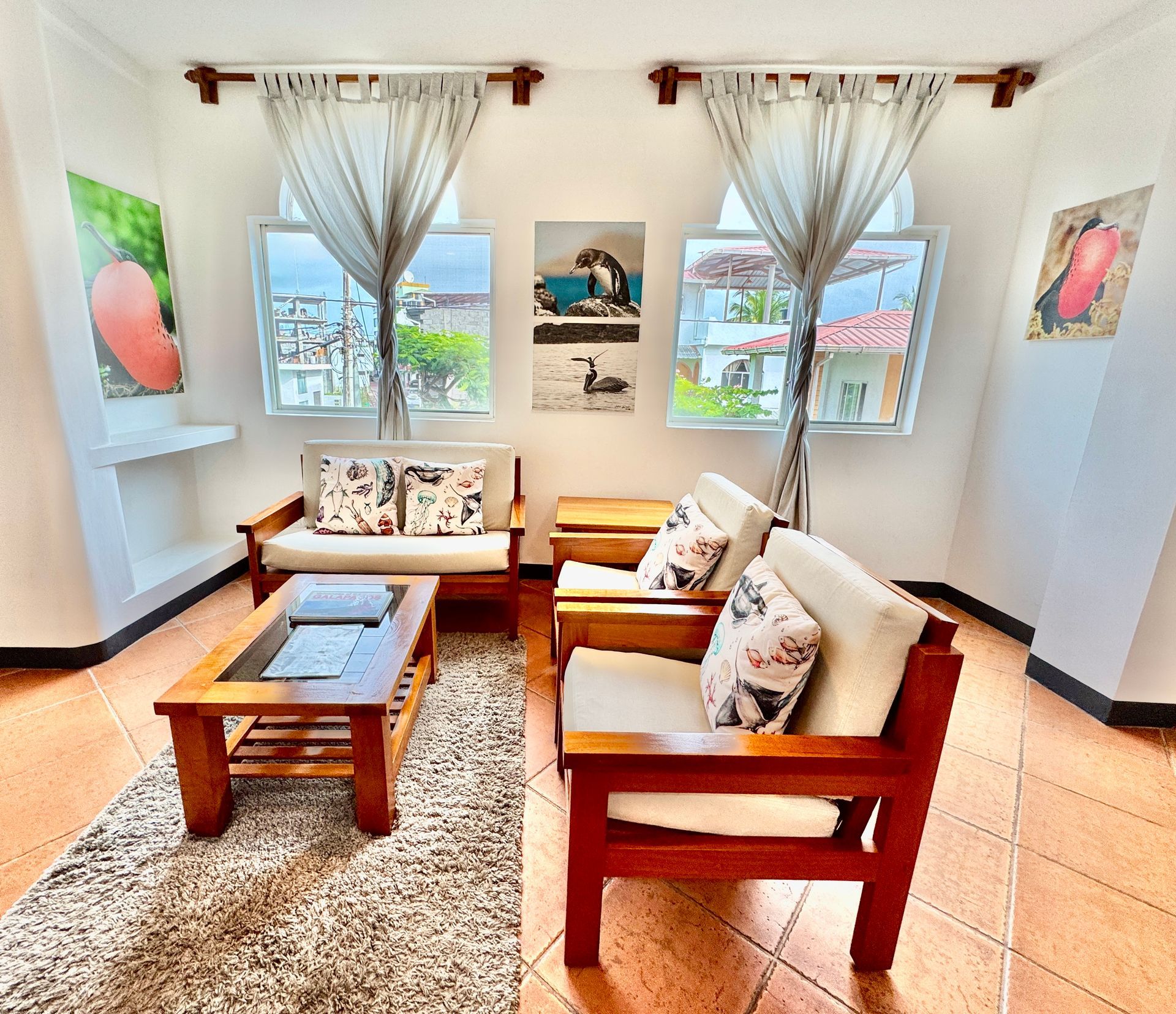 A well-lit living room with wooden furniture, beige cushions, and art on the walls. Two windows offer a view of the outside.