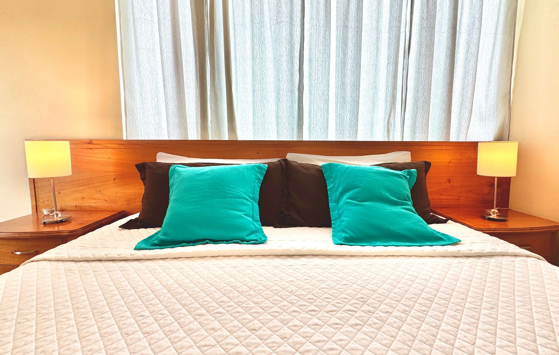 A bed in a bedroom with teal and brown pillows, matching nightstands, and a window with curtains.