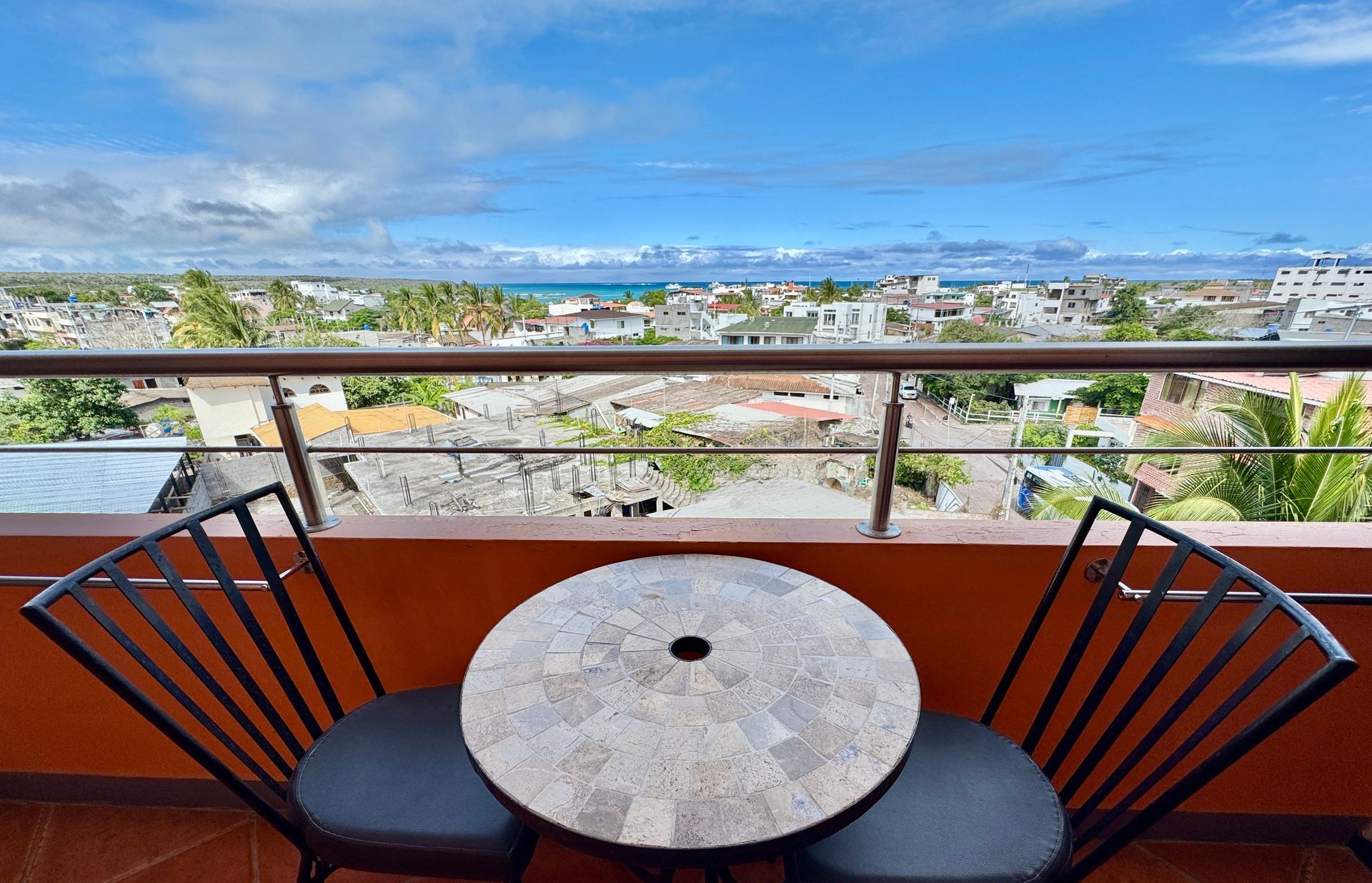 A balcony with a round table and chairs overlooking a city and the ocean under a blue sky.