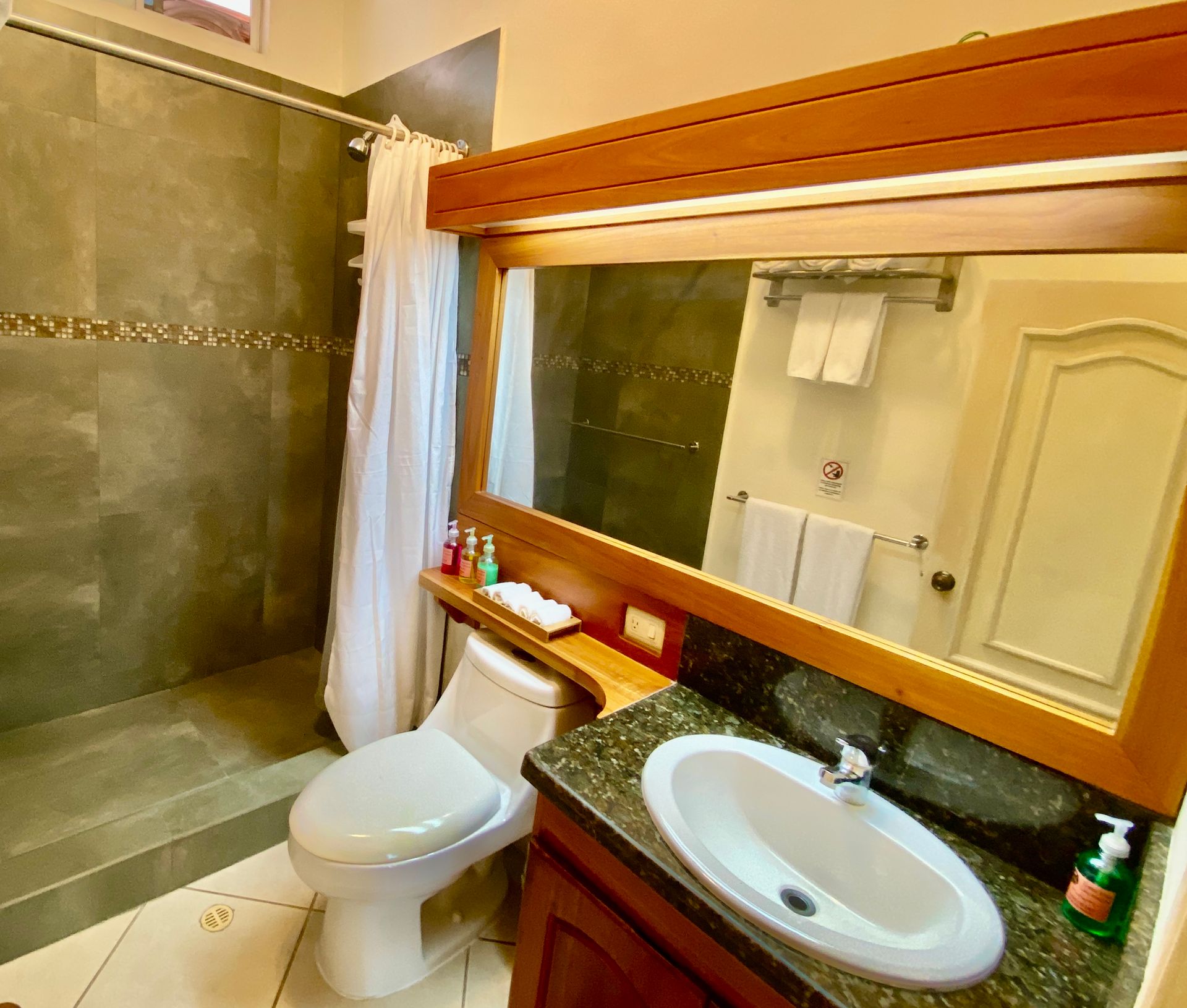Bathroom with a toilet, sink, shower, and large mirror. Wooden accents, grey tile, and white towels.