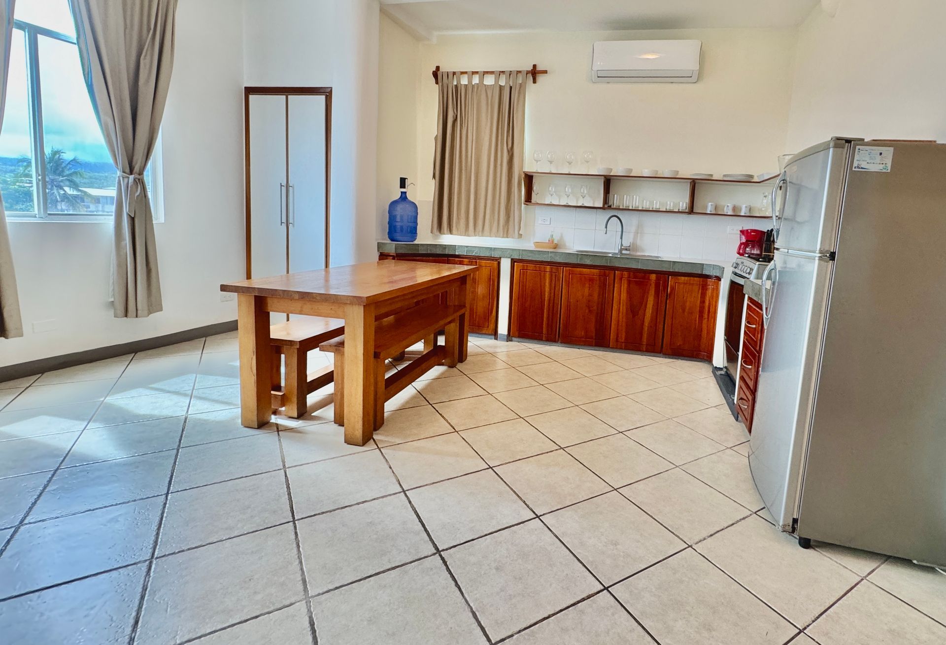 A bright kitchen with a wooden table, cabinets, and a refrigerator. Natural light streams through a window.