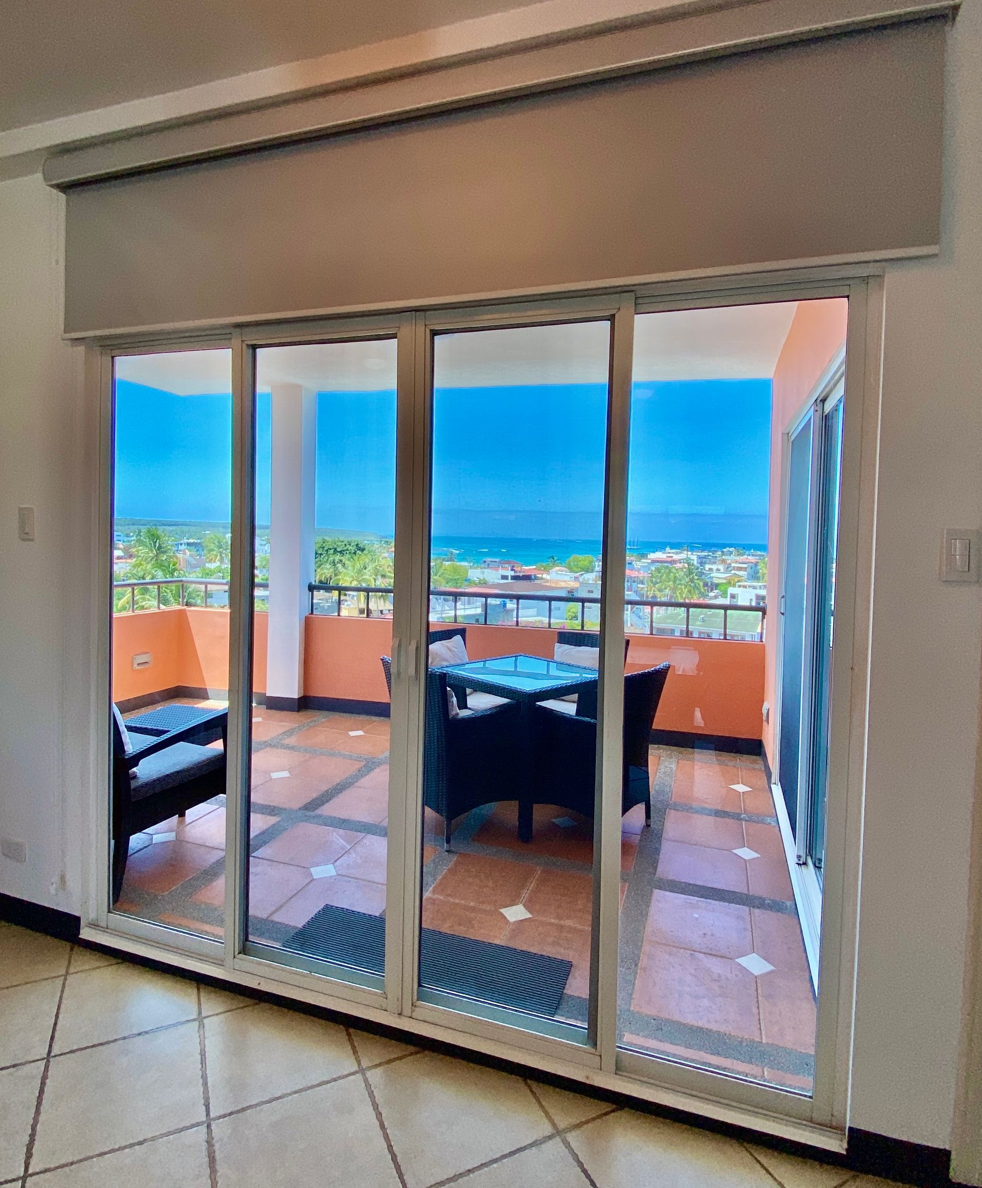 Sliding glass doors open to a balcony with a table and chairs, overlooking a blue ocean and sky.