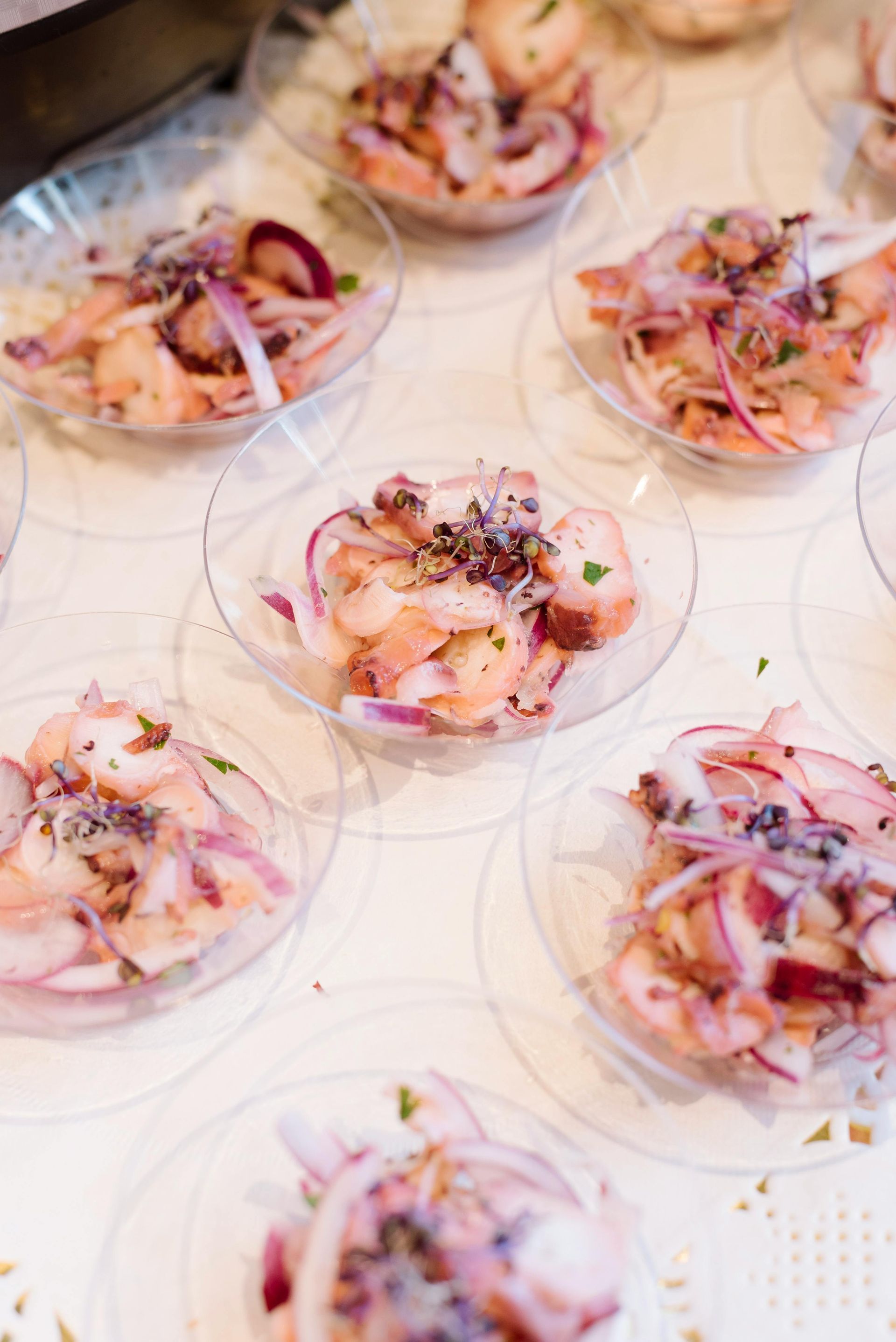 Small, clear plastic bowls filled with ceviche, each topped with red onion and herbs, arranged on a white surface.
