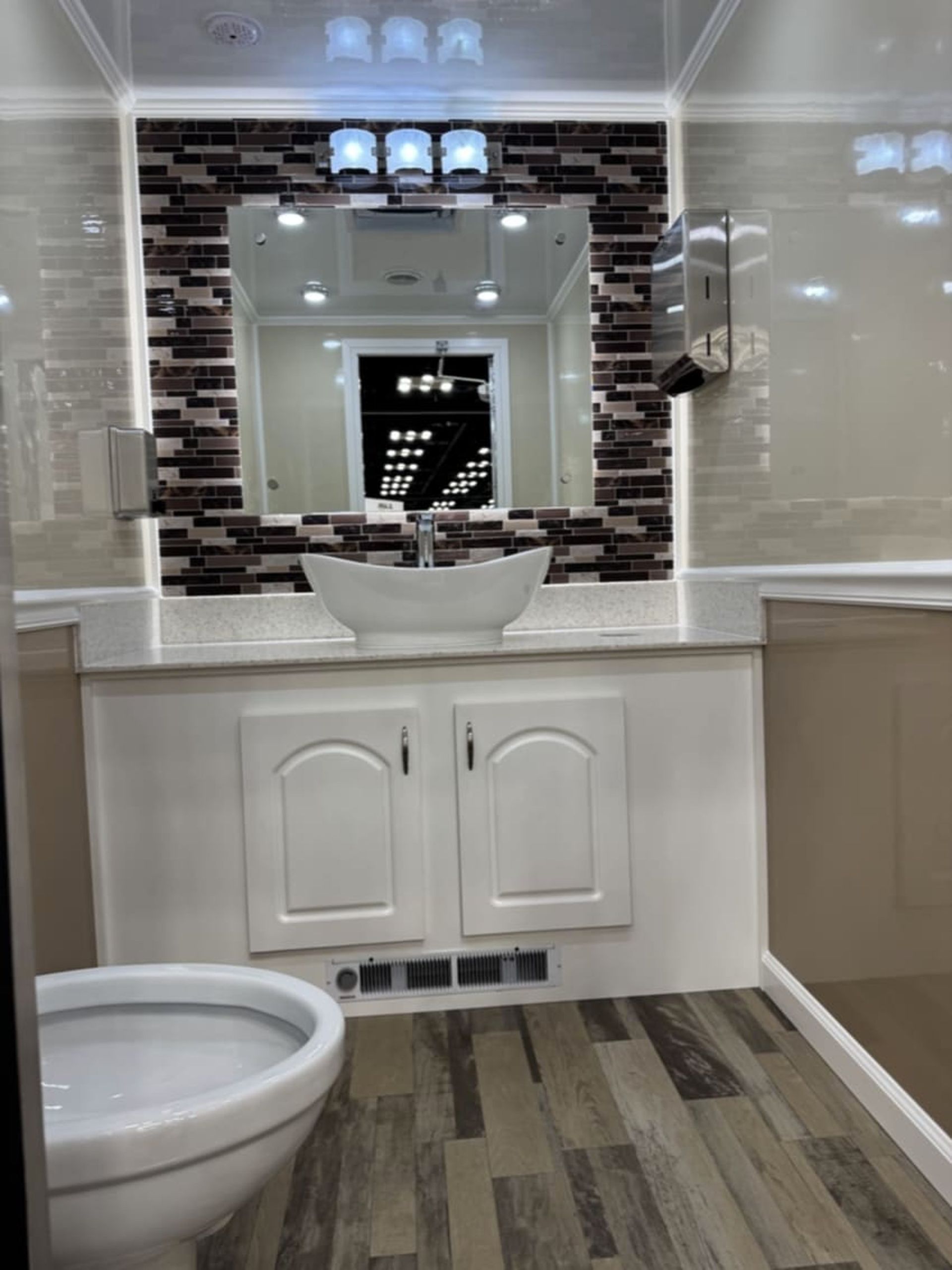Interior of a luxury portable restroom: white sink, mirror with brown tile, toilet, and wood-look floor.