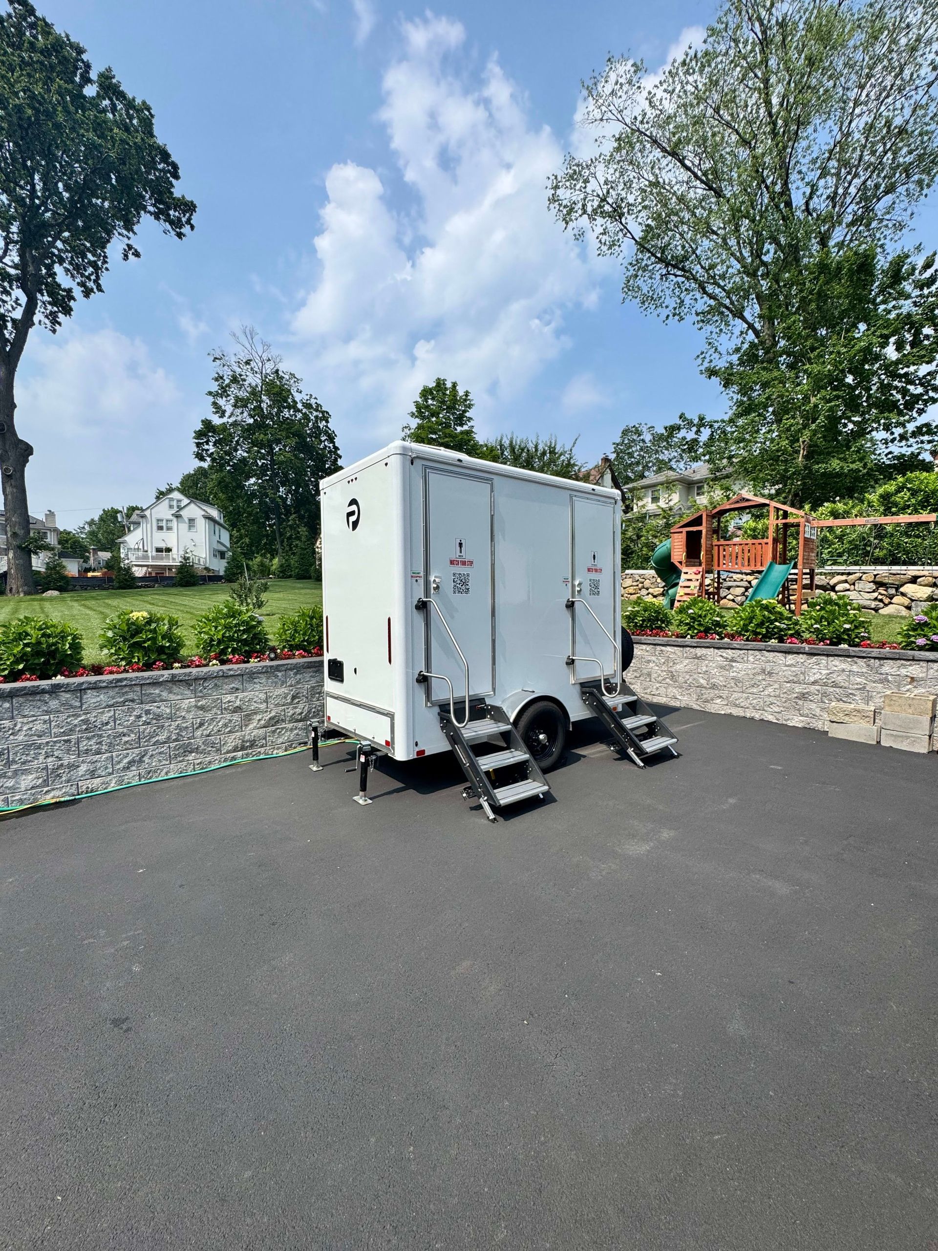 White portable restroom trailer on blacktop driveway, surrounded by green landscaping under a blue sky.