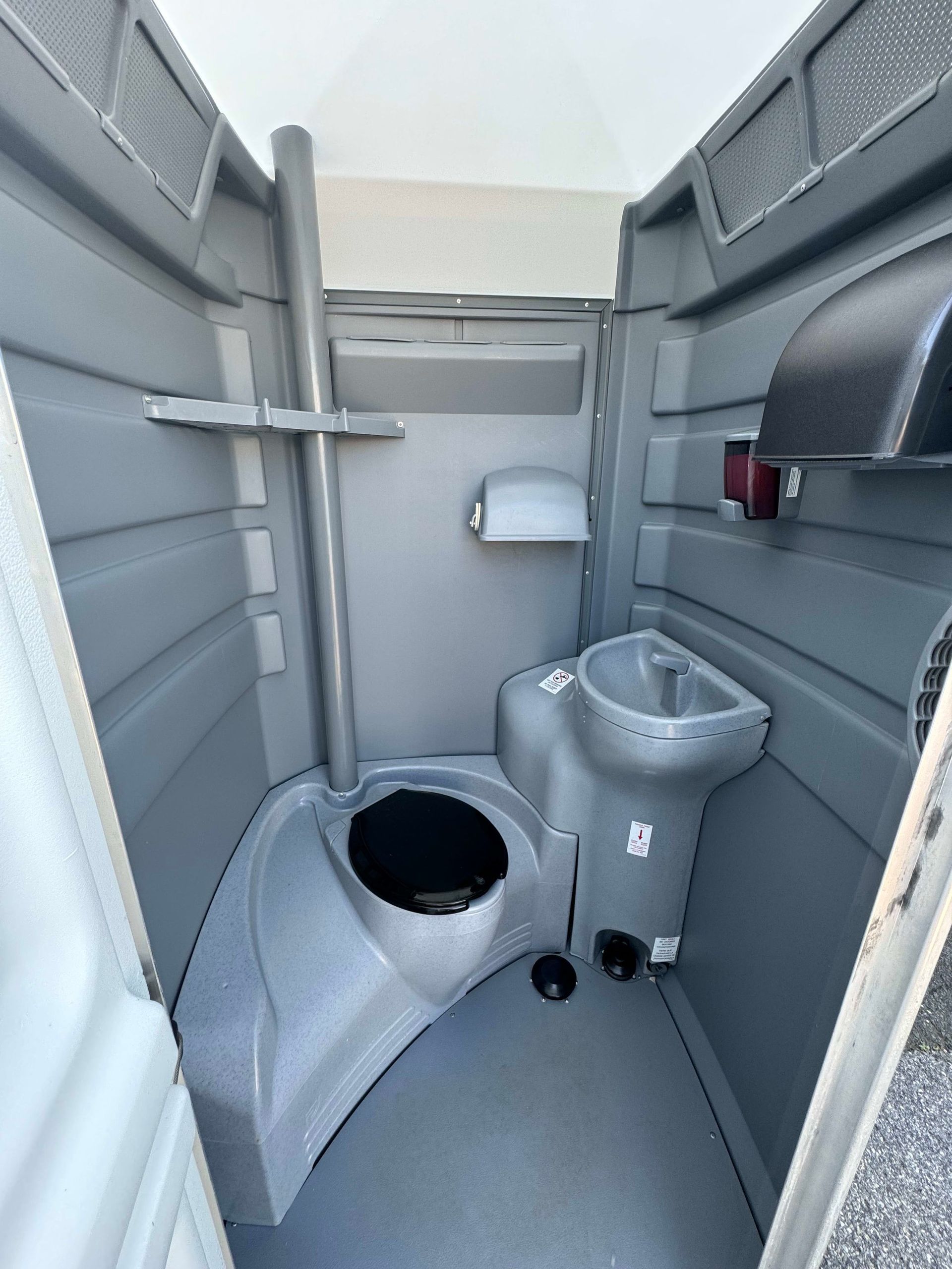 Interior of a portable toilet. Gray plastic walls, toilet, and sink are visible. Door is open.