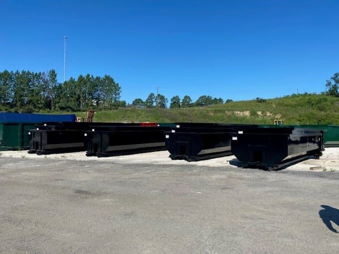 Four black dumpsters sit outside under a clear blue sky. Trees and a tall pole are in the background.