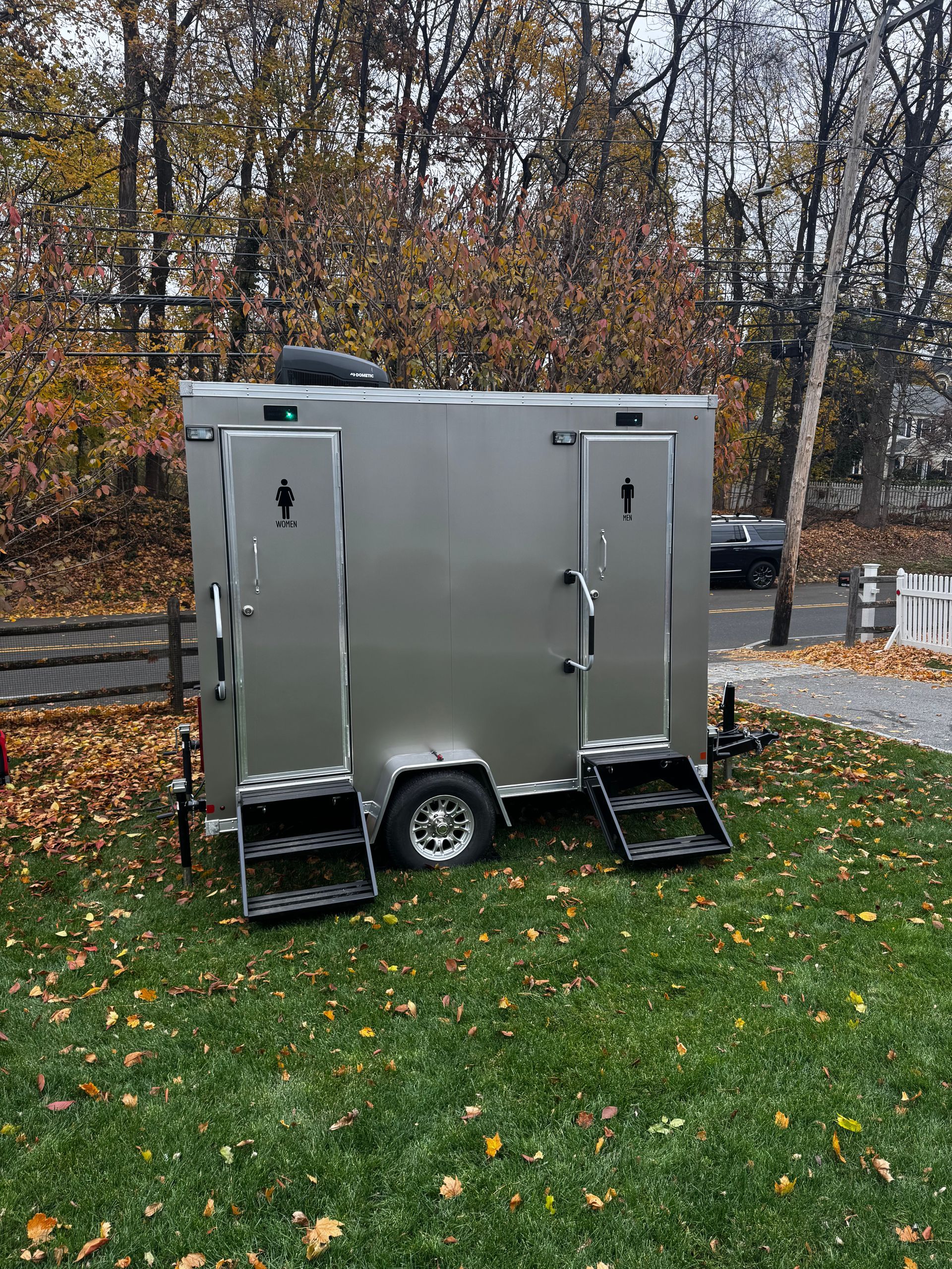 White portable restroom trailer with steps, parked on driveway next to gray house.