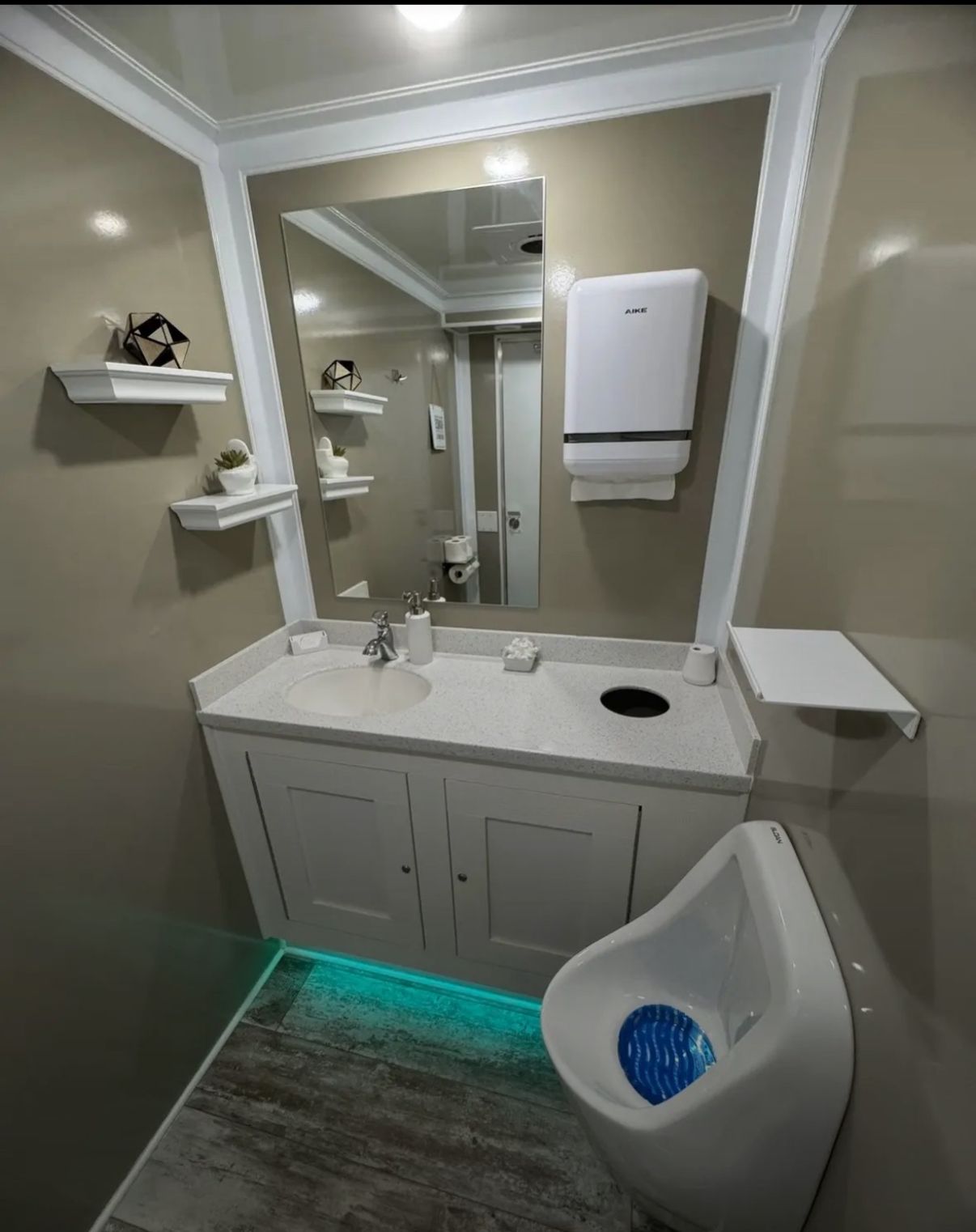 A modern bathroom featuring a white vanity with a mirror, a wall-mounted paper towel dispenser, and a white urinal.