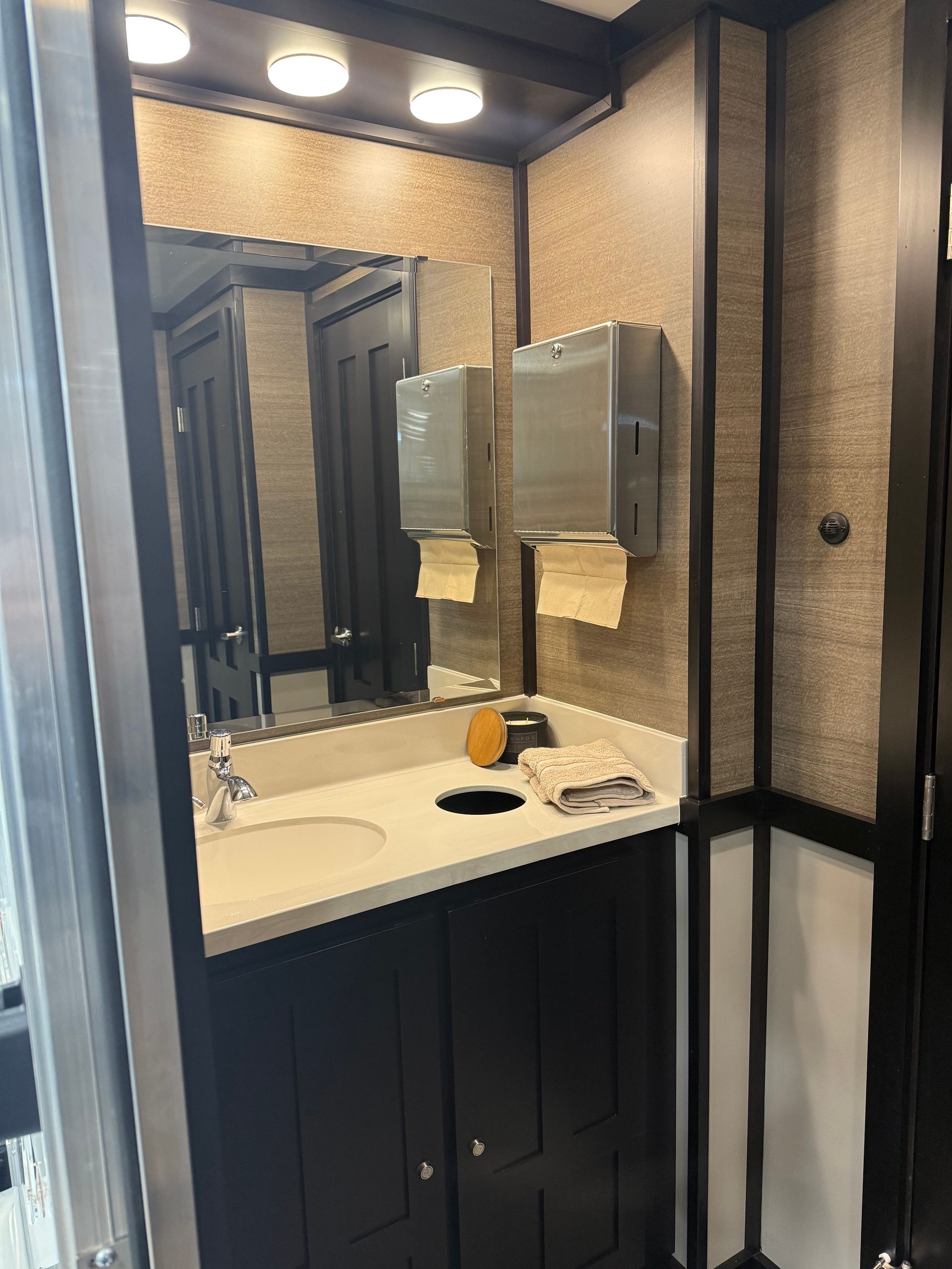 A restroom vanity with a white counter, black cabinet, sink, faucet, mirror, and stainless steel paper towel dispenser.
