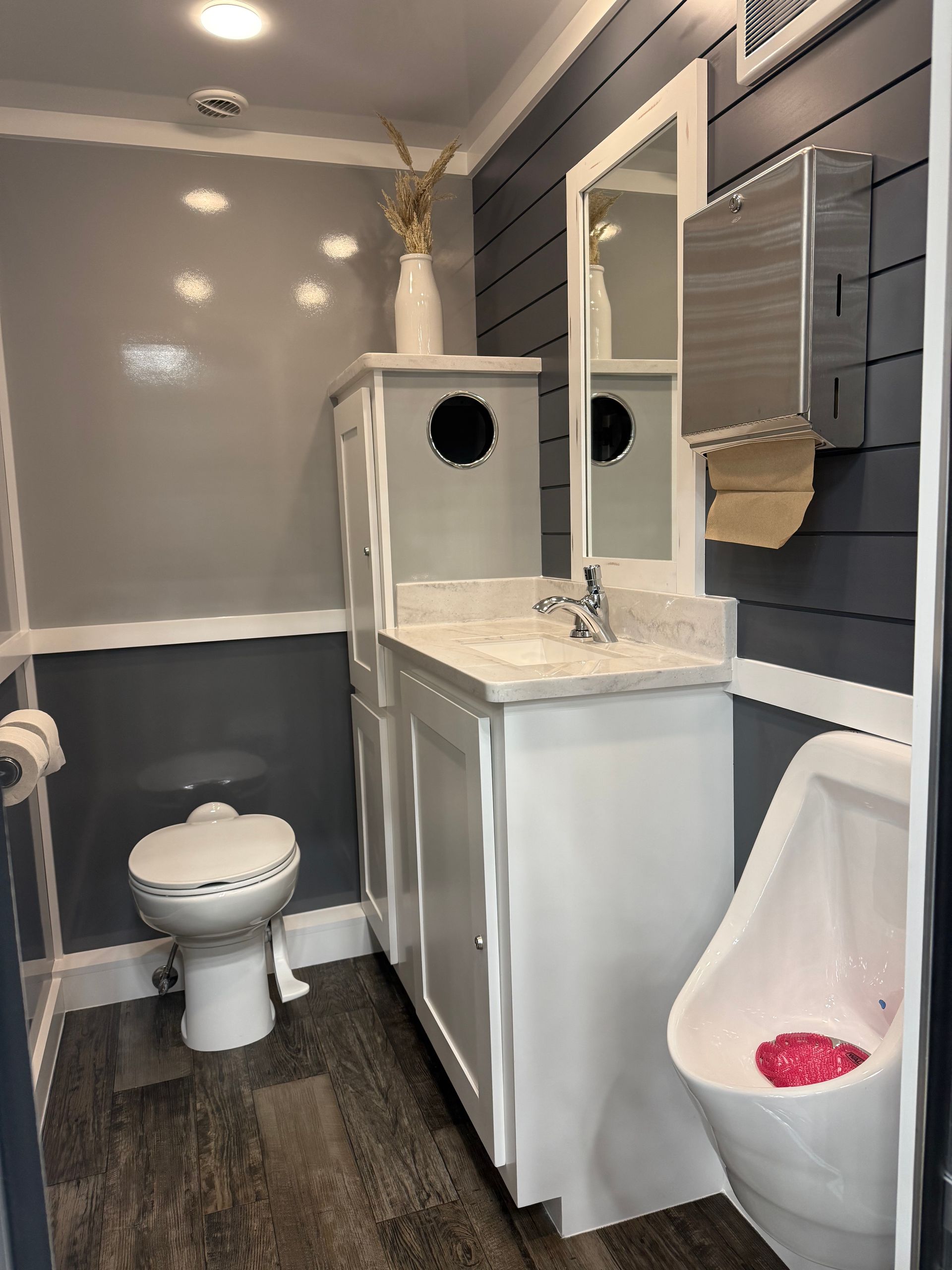 Small restroom featuring a white toilet, white vanity with mirror, and urinal against gray shiplap walls and wood flooring.