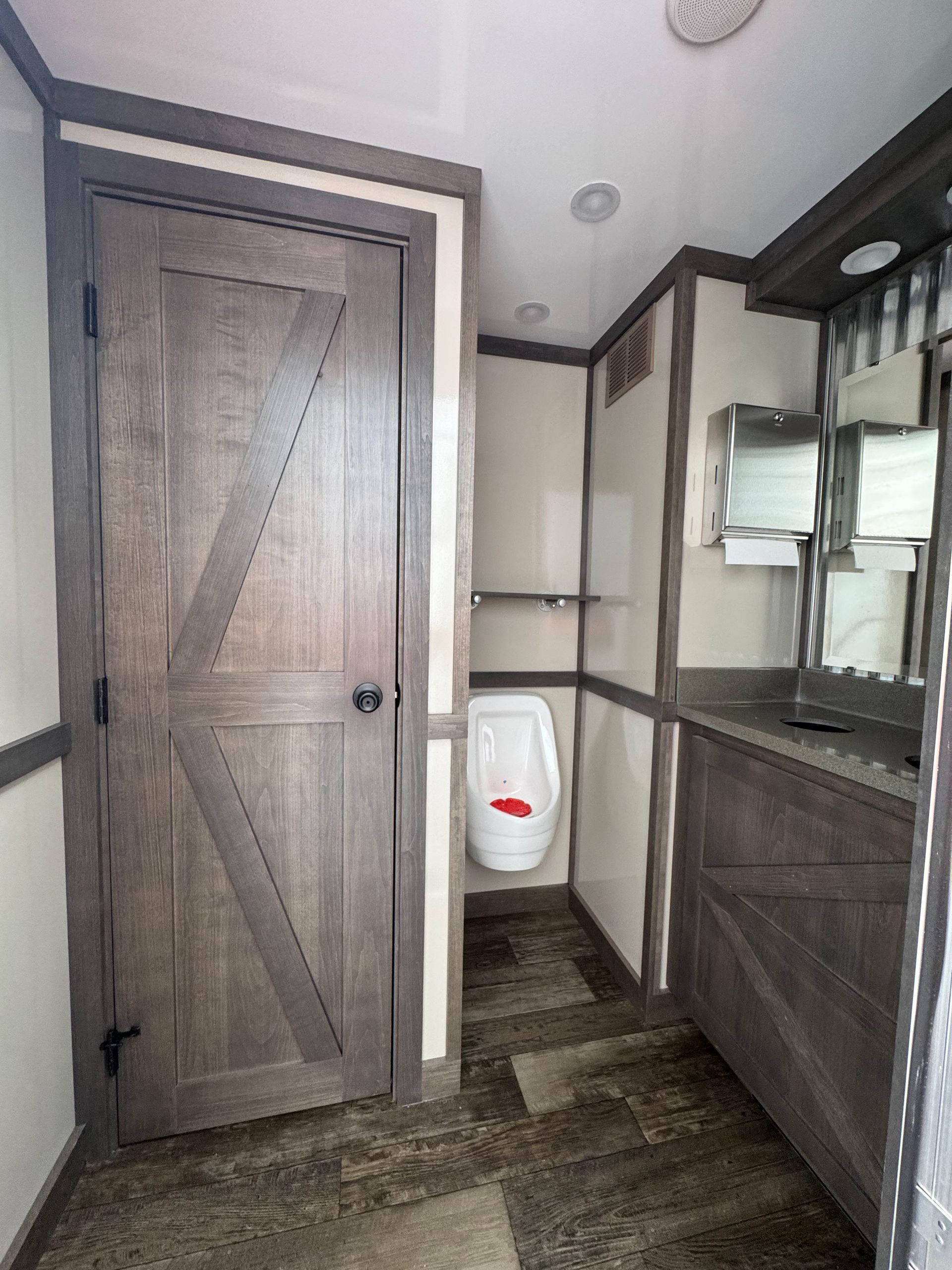 Interior of a portable restroom with a wooden door, urinal, sink, and neutral color scheme.