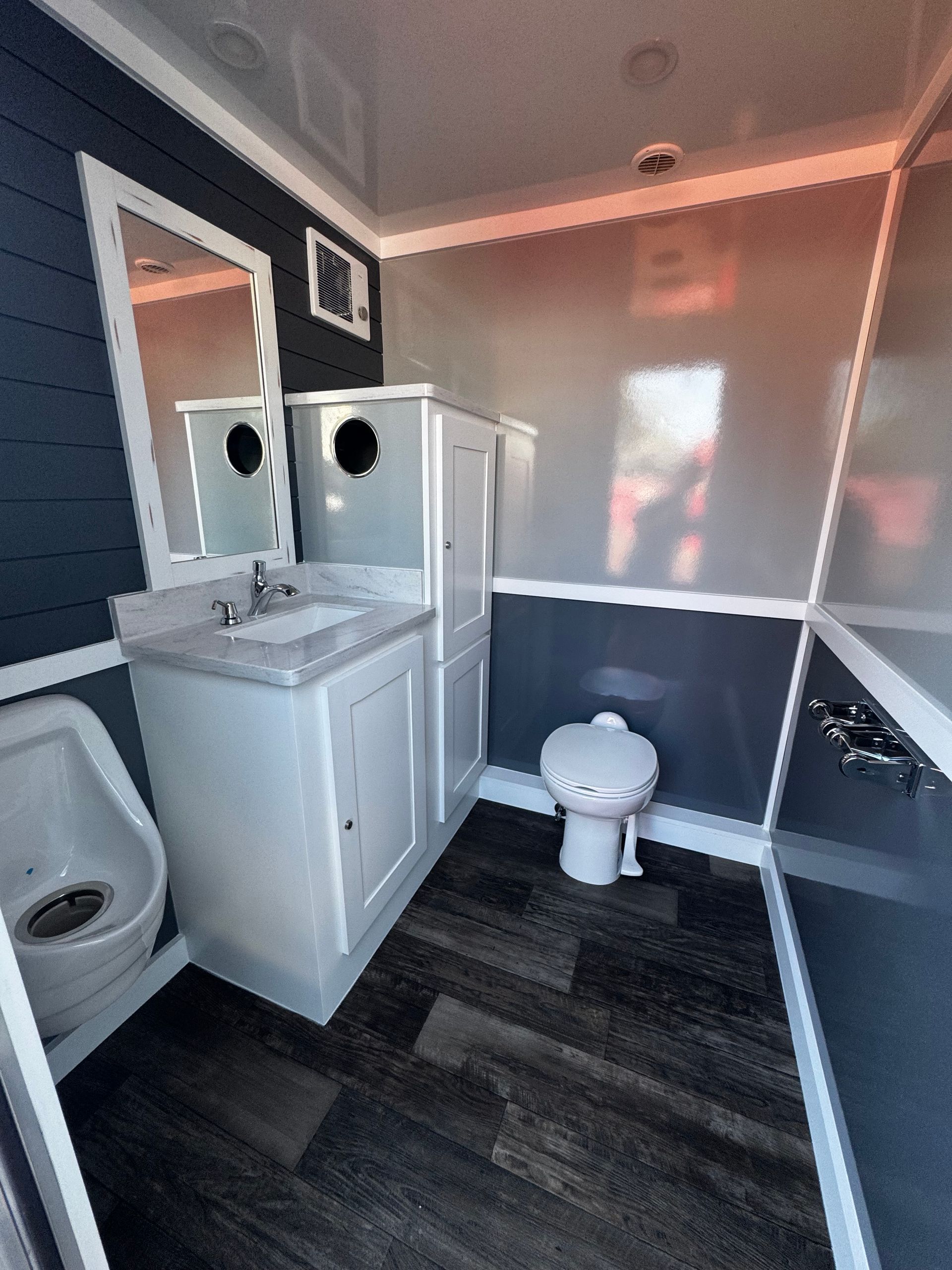 Interior of a portable restroom with a sink, toilet, and urinal. Blue walls and dark wood-look floor.