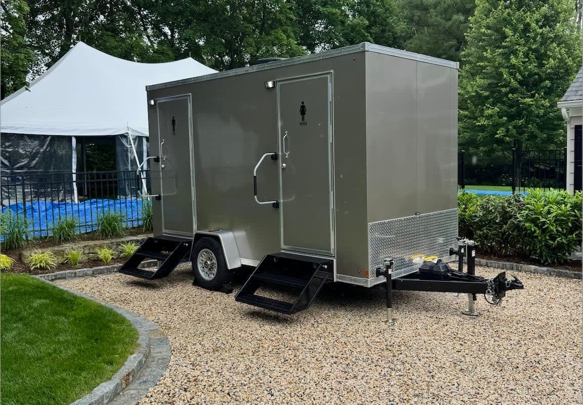 Portable restroom trailer with two doors, gray exterior, gravel base, steps. Tent in background.