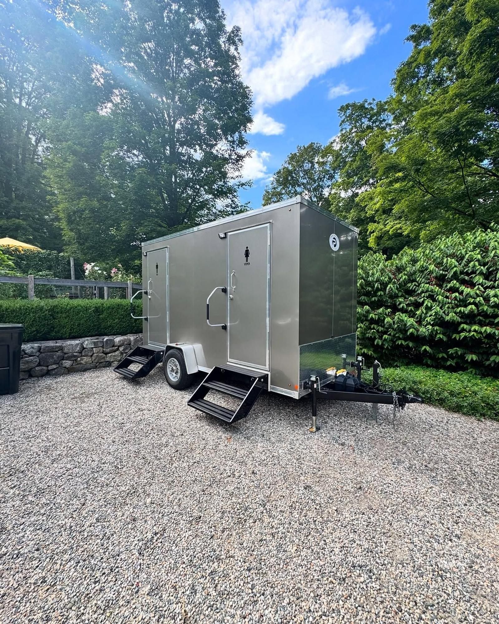A silver portable restroom trailer on gravel, steps in front, surrounded by greenery and a blue sky.