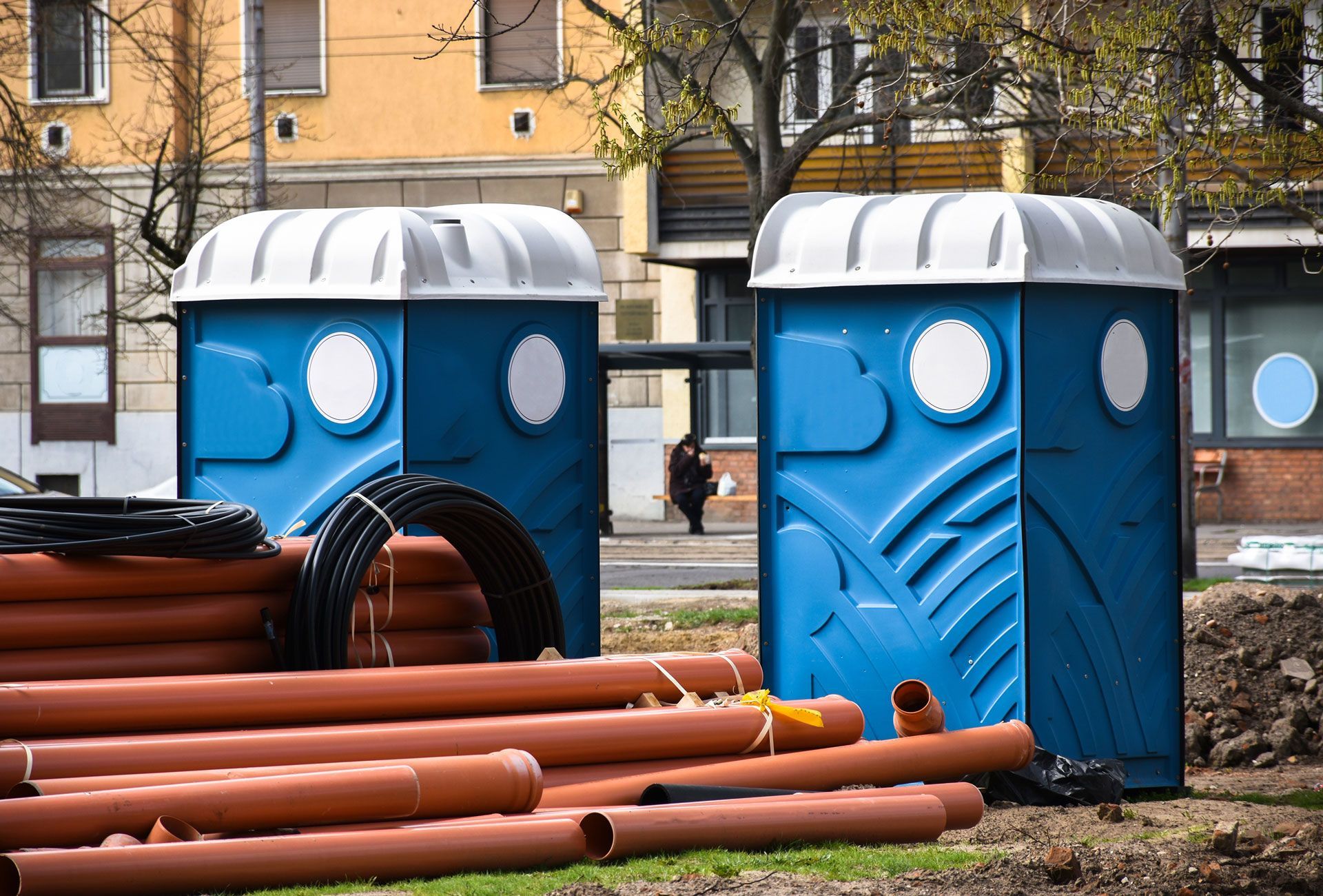 Two blue porta-potties, brown pipes, and black coils on construction site, with a person in the background.