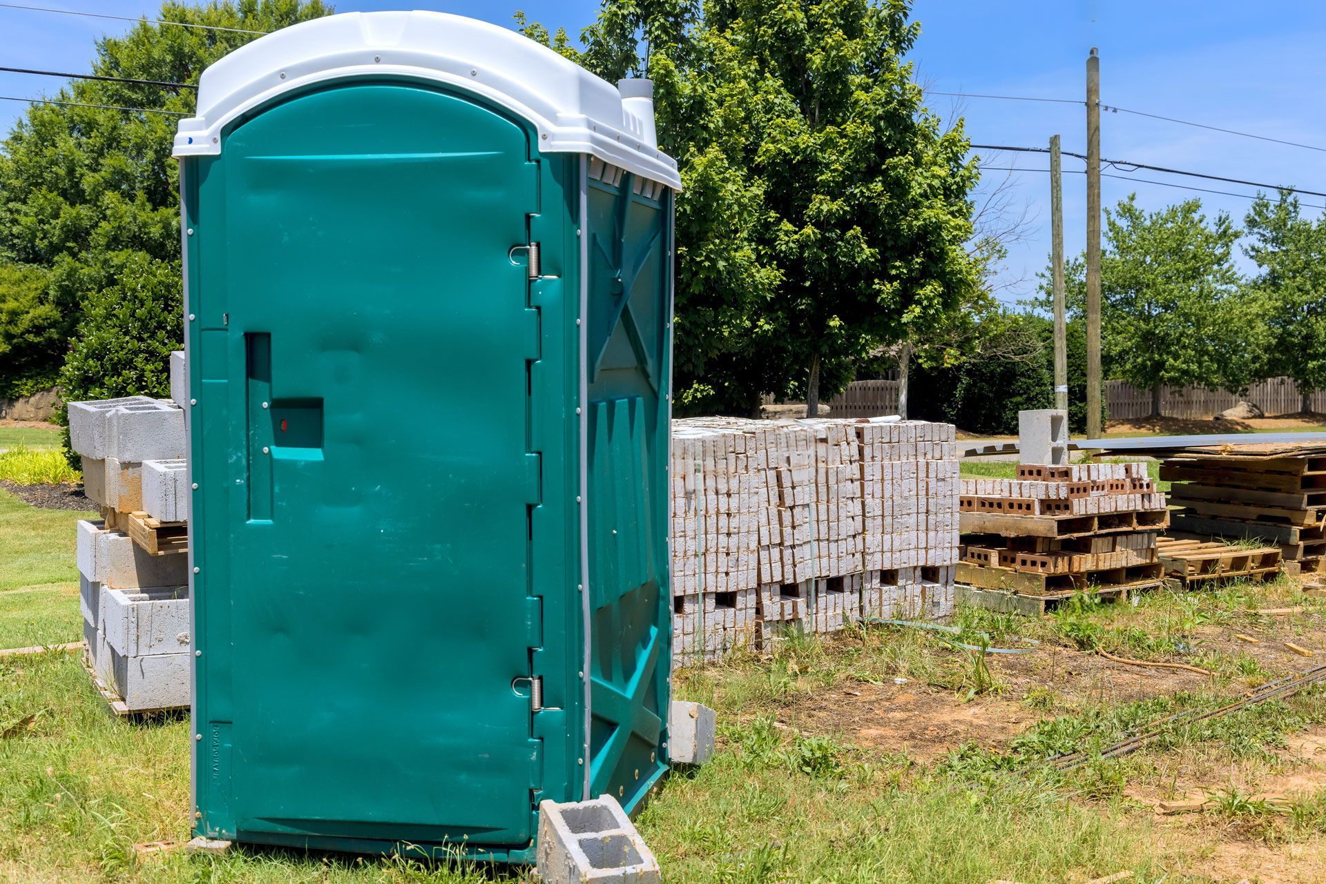 Green porta-potty with white top, outside with stacked cinder blocks, green trees and sunny sky in the background.