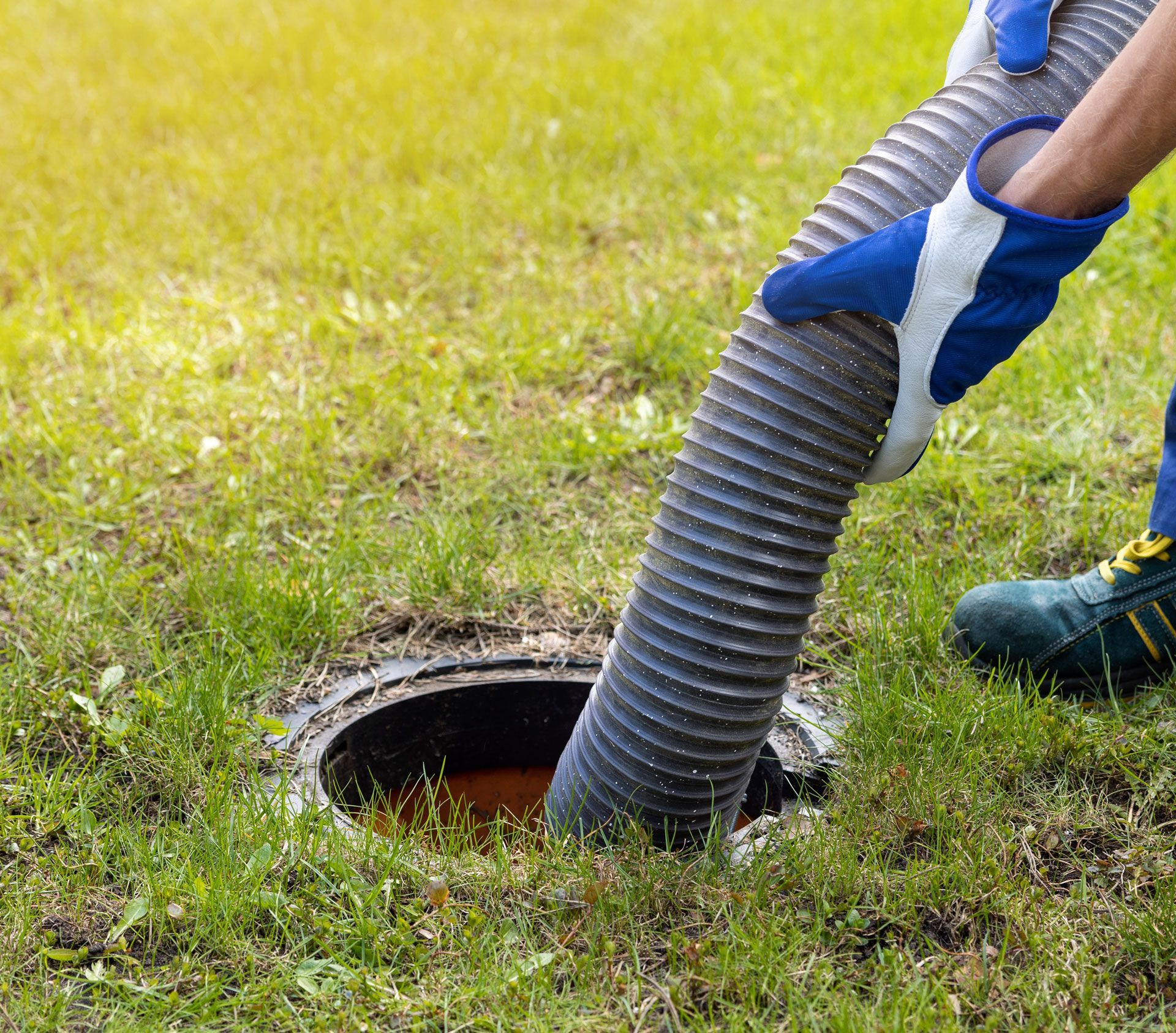 A worker wearing protective gloves uses a large vacuum hose to clear out an underground septic tank.