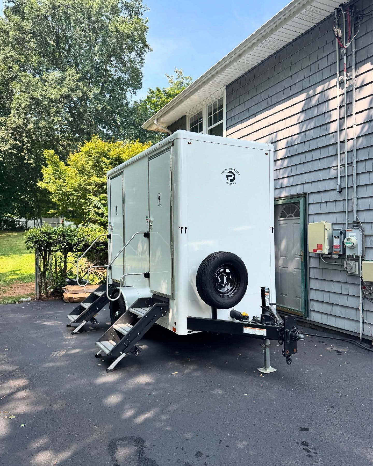 White portable restroom trailer with steps, parked on driveway next to gray house.