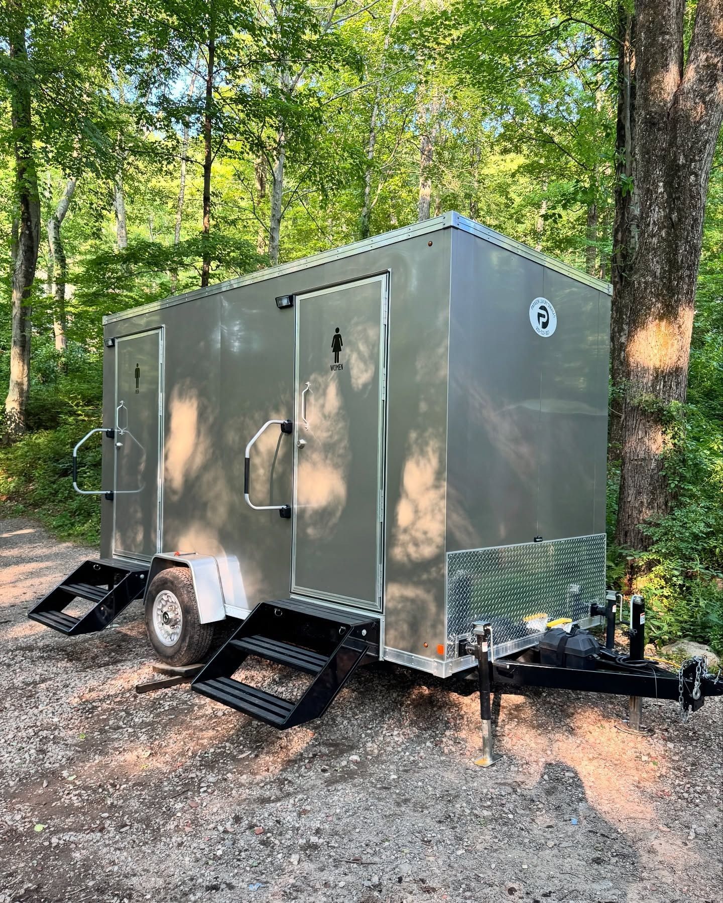 Portable restroom trailer with two stalls, silver metal, set outdoors on gravel path, black steps.