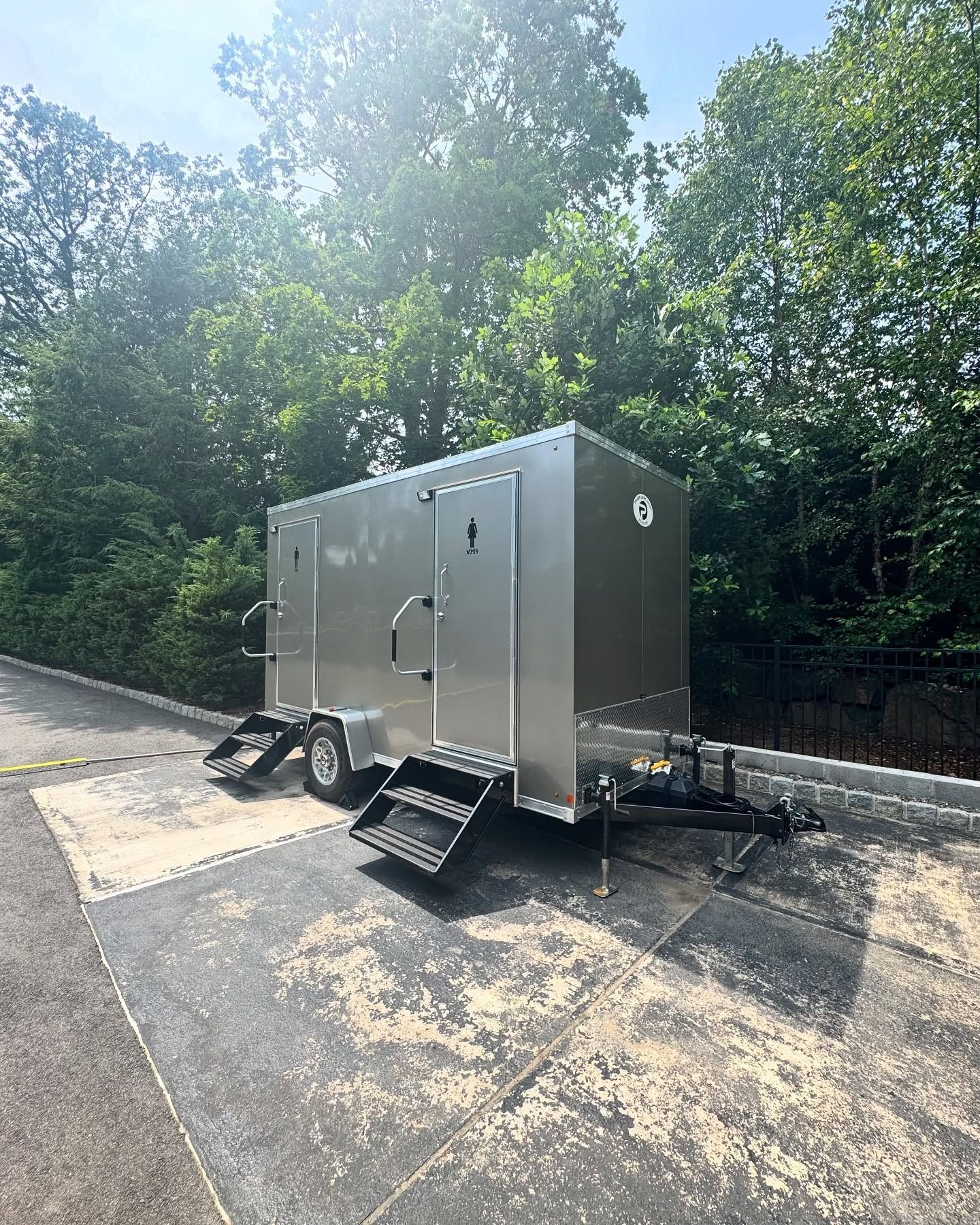 Silver portable restroom trailer with steps parked on asphalt, trees in background.