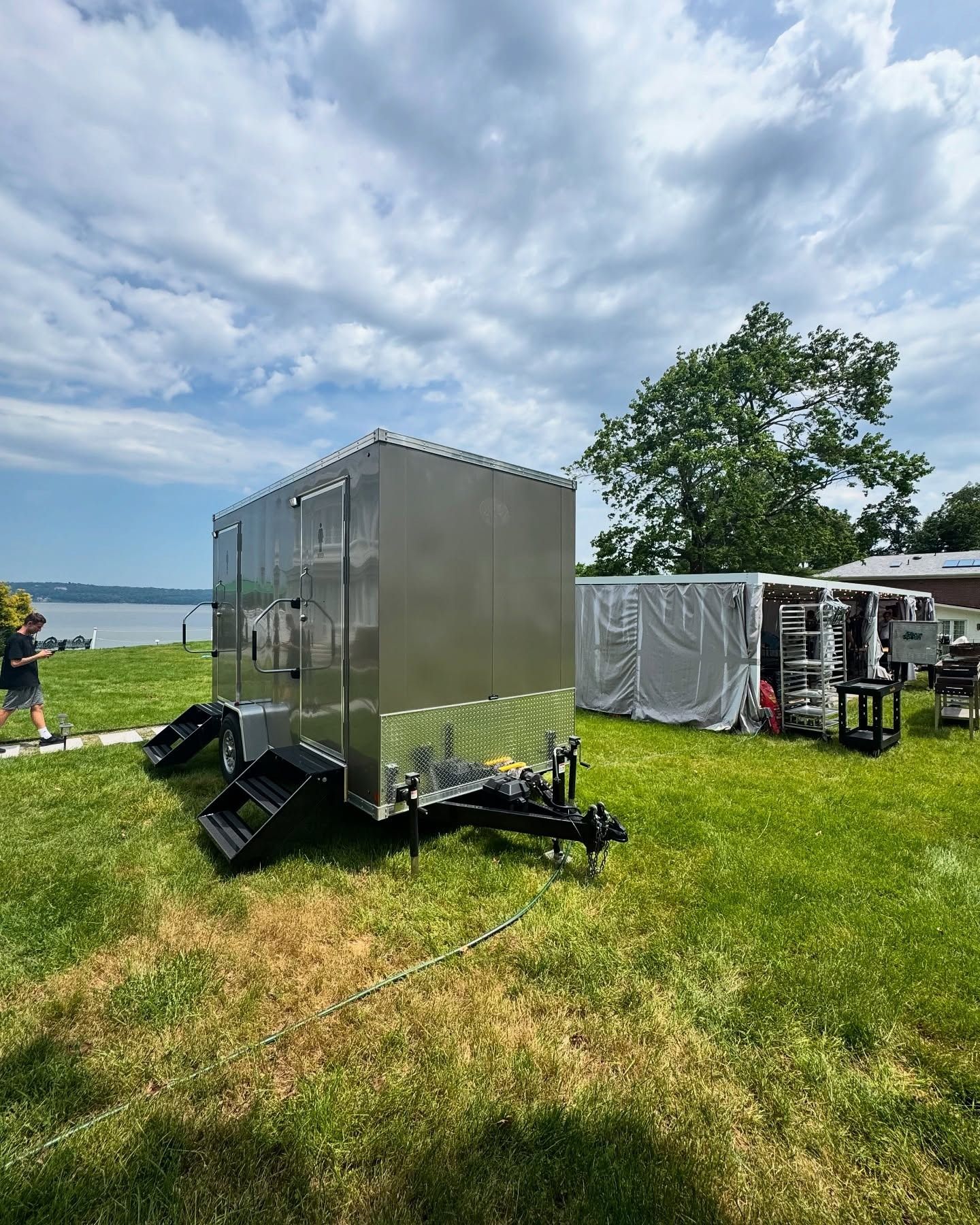 A portable restroom trailer and a white tent are set up on a grassy lawn near a body of water under a cloudy sky.