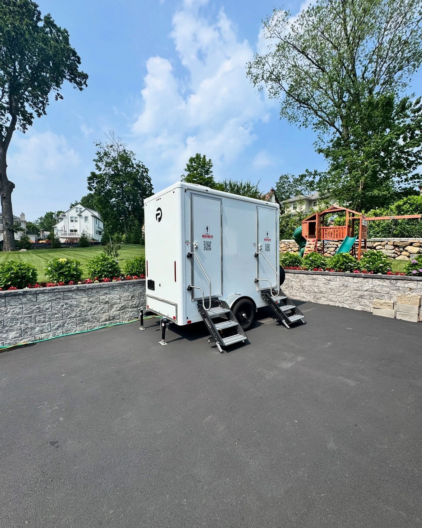 White portable restroom trailer on black pavement with stairs, against a background of green trees and a blue sky.