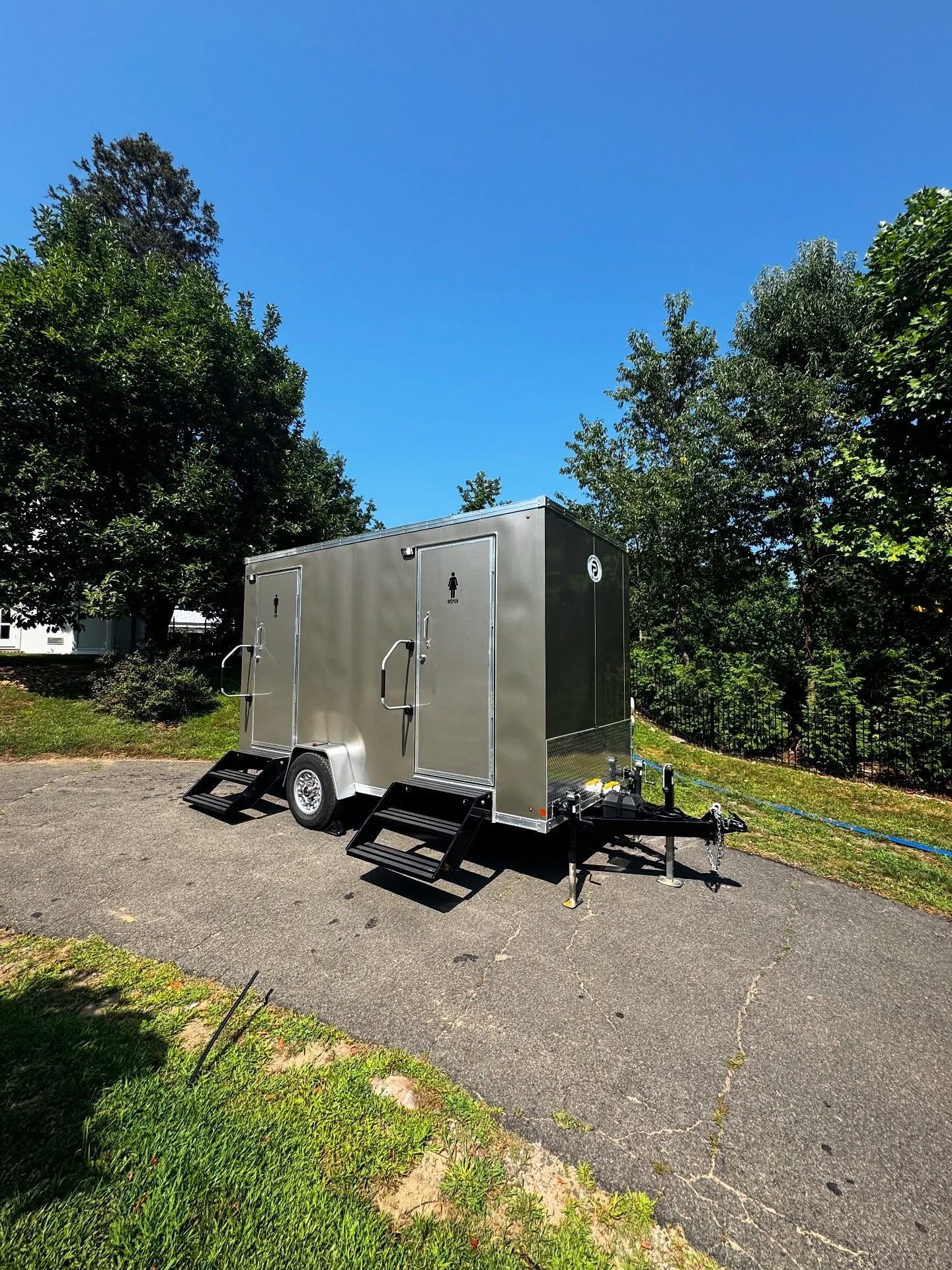 A stainless steel portable restroom trailer on a paved driveway, steps leading up to the entrance.