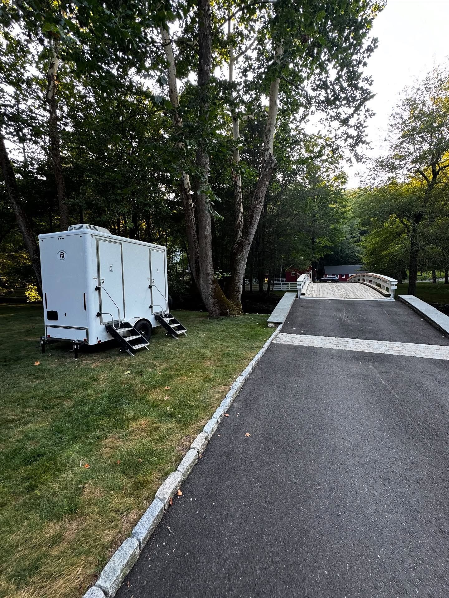 White portable restrooms next to a paved path with a small bridge, set in a green grassy area with trees.