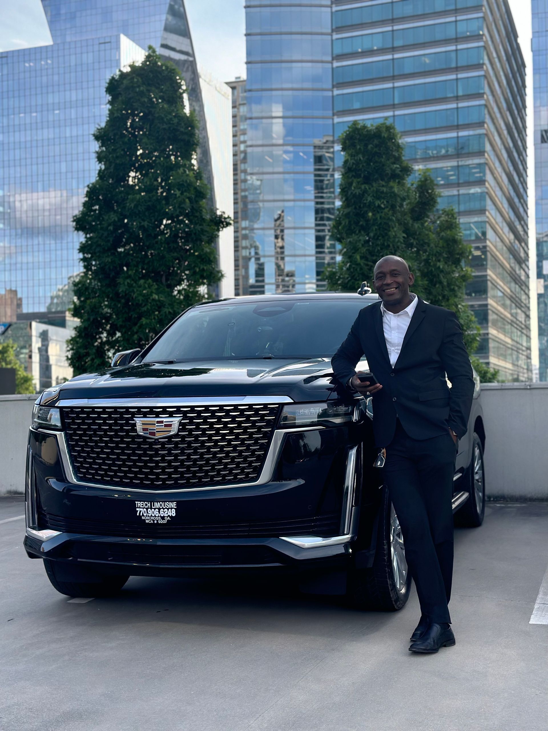 Man in suit poses next to a black Cadillac Escalade in front of skyscrapers.
