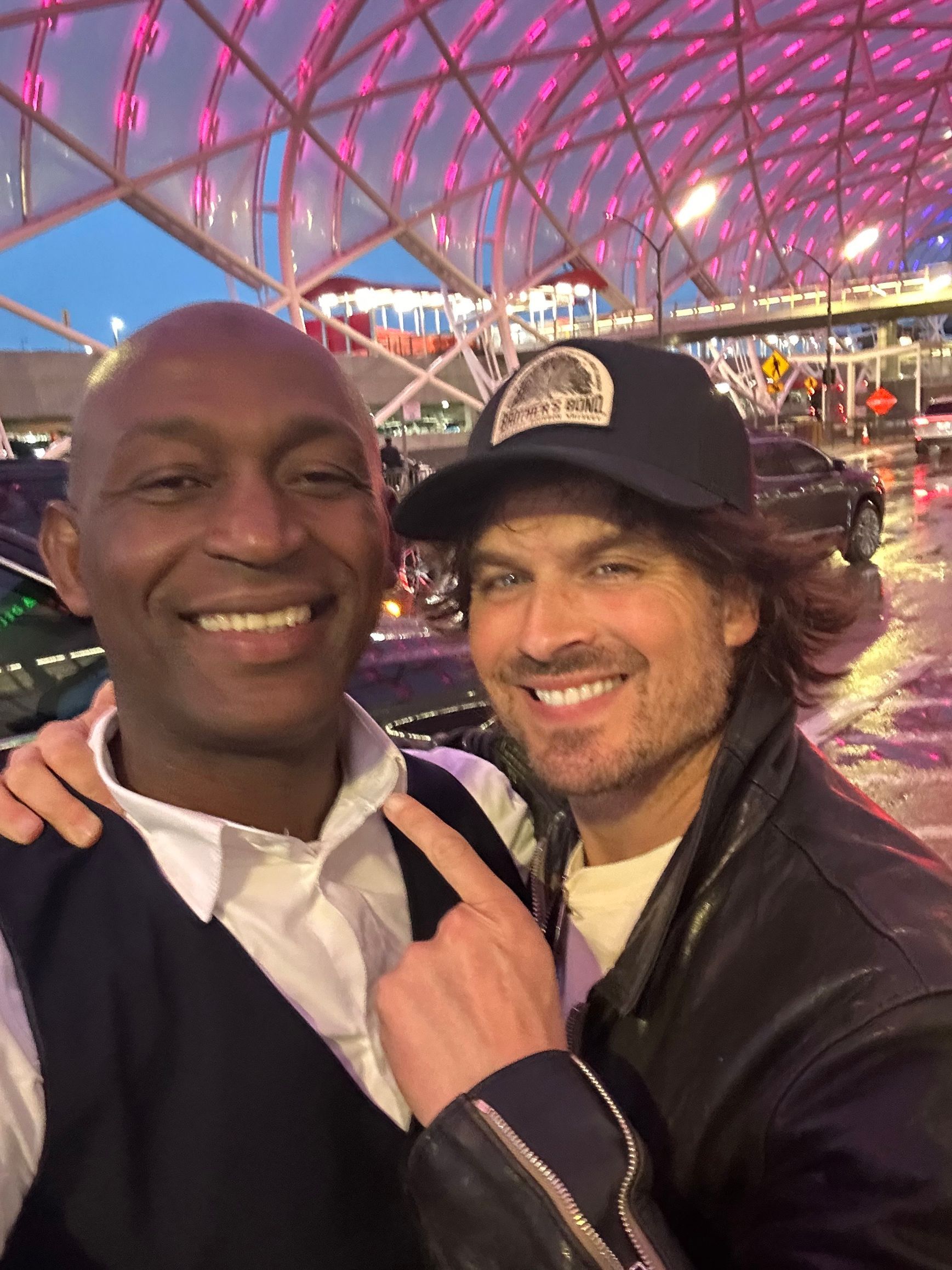 Two men smile for a selfie at an airport. One points. One wears a cap and leather jacket. Night.