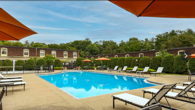 Swimming pool with lounge chairs and orange umbrellas, surrounded by hedges and buildings under a blue sky.